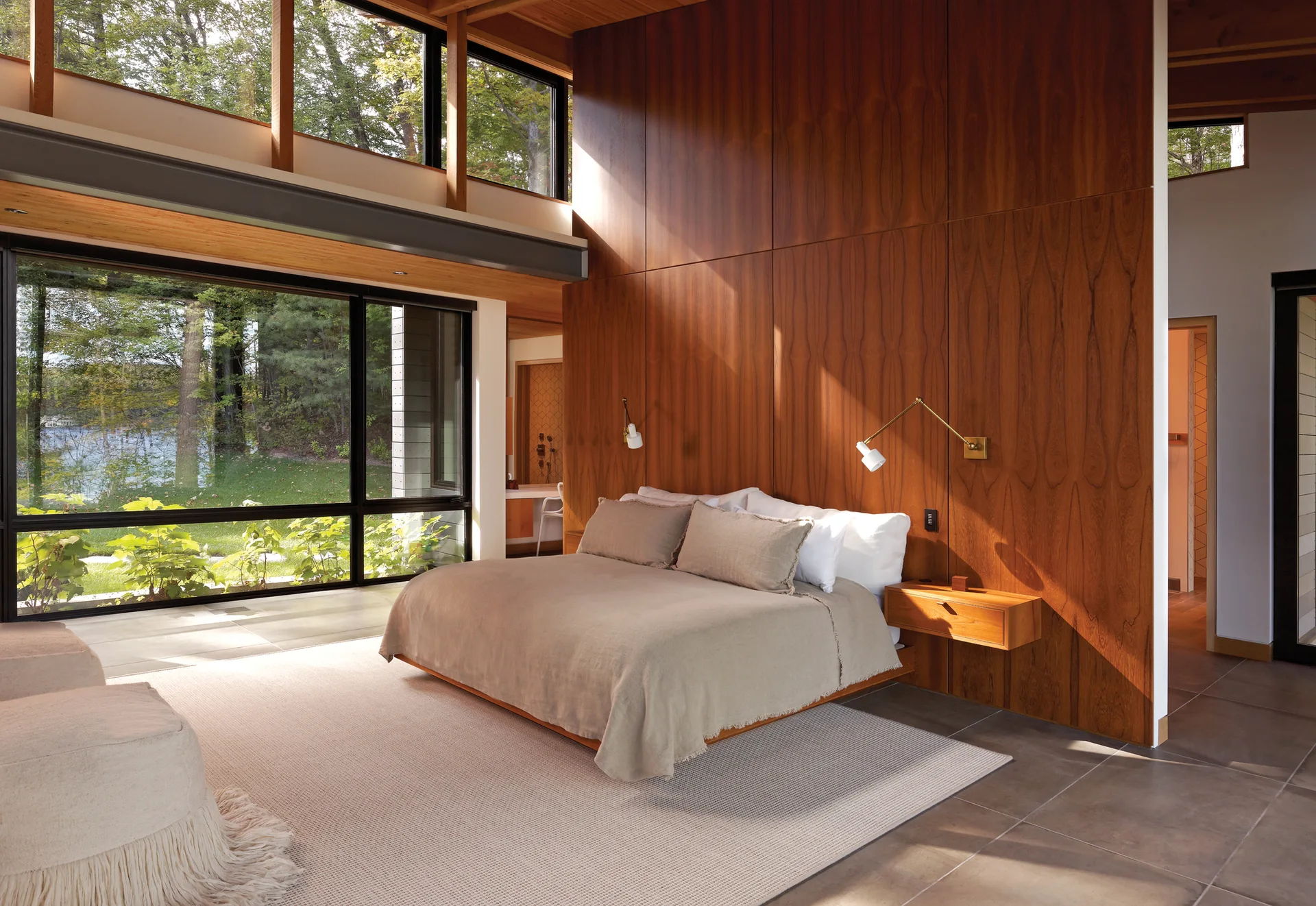 A modern bedroom features a low bed against a tall wood-paneled accent wall, flanked by a floating nightstand and large glass windows.