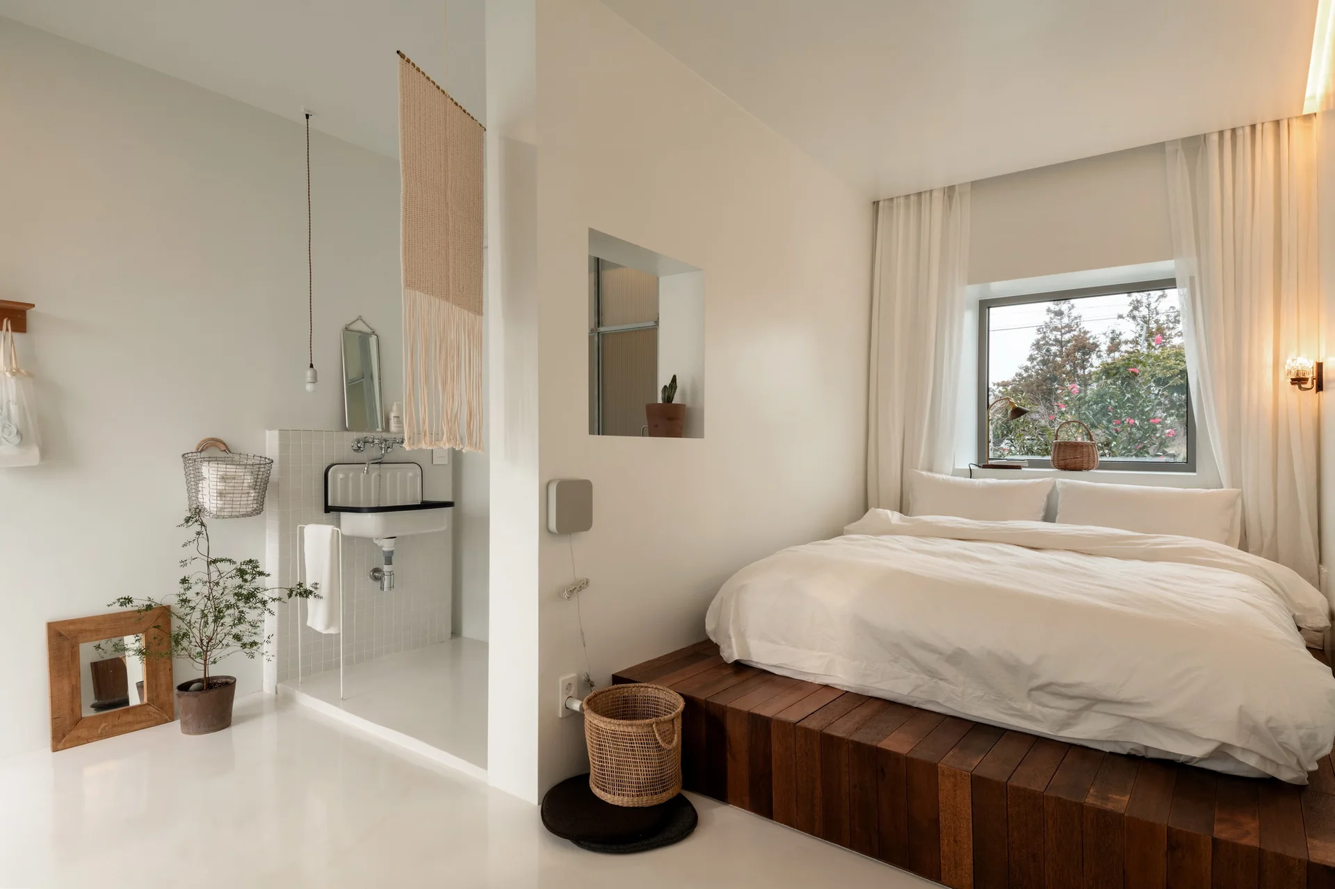 A minimalist bedroom features a dark wood platform bed with white linens next to an open bathroom nook with a tiled wall and sink.
