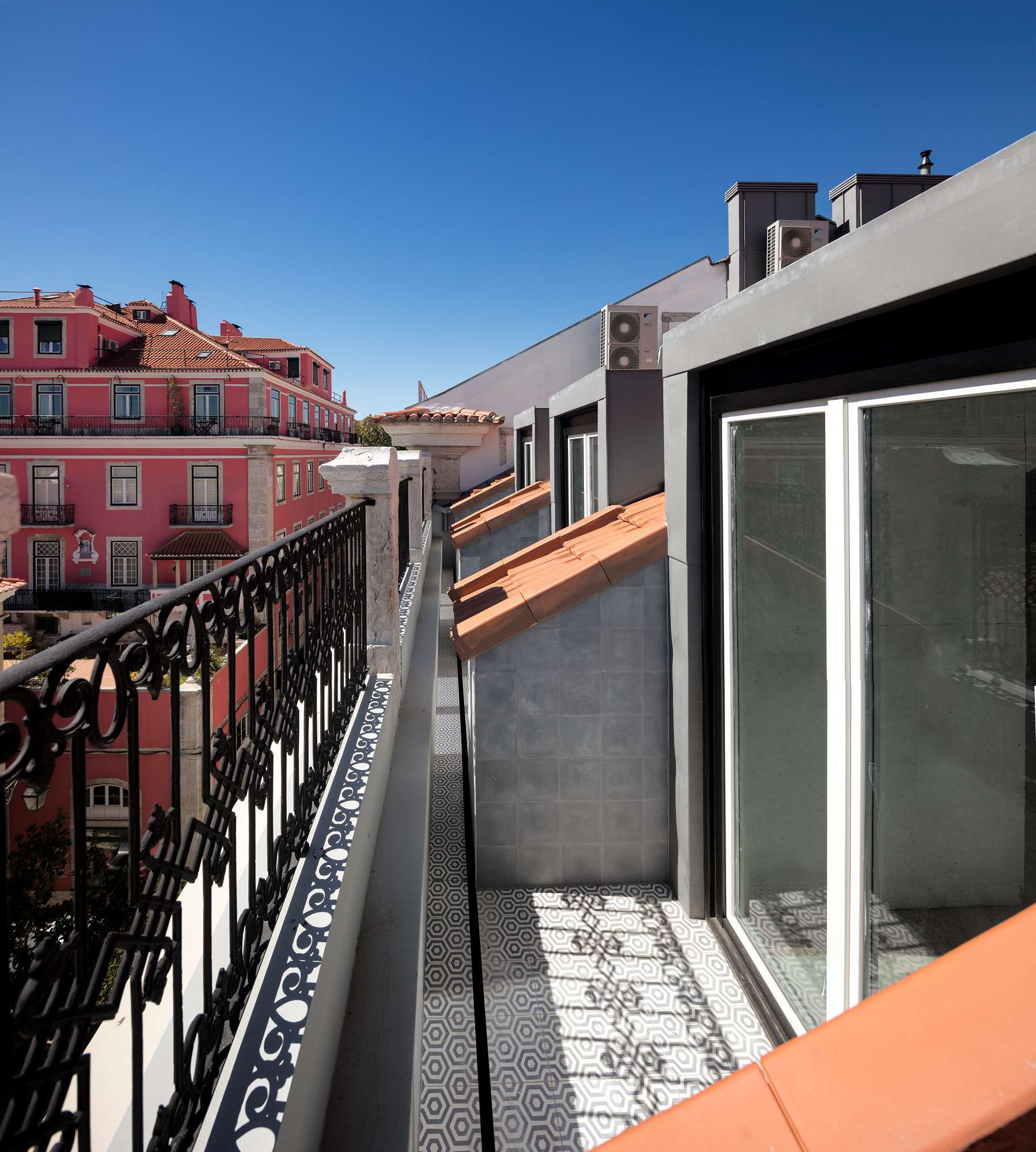 A sunny, narrow outdoor balcony featuring grey and white geometric hexagonal floor tiles, a black wrought iron railing, and a view of a pink building.