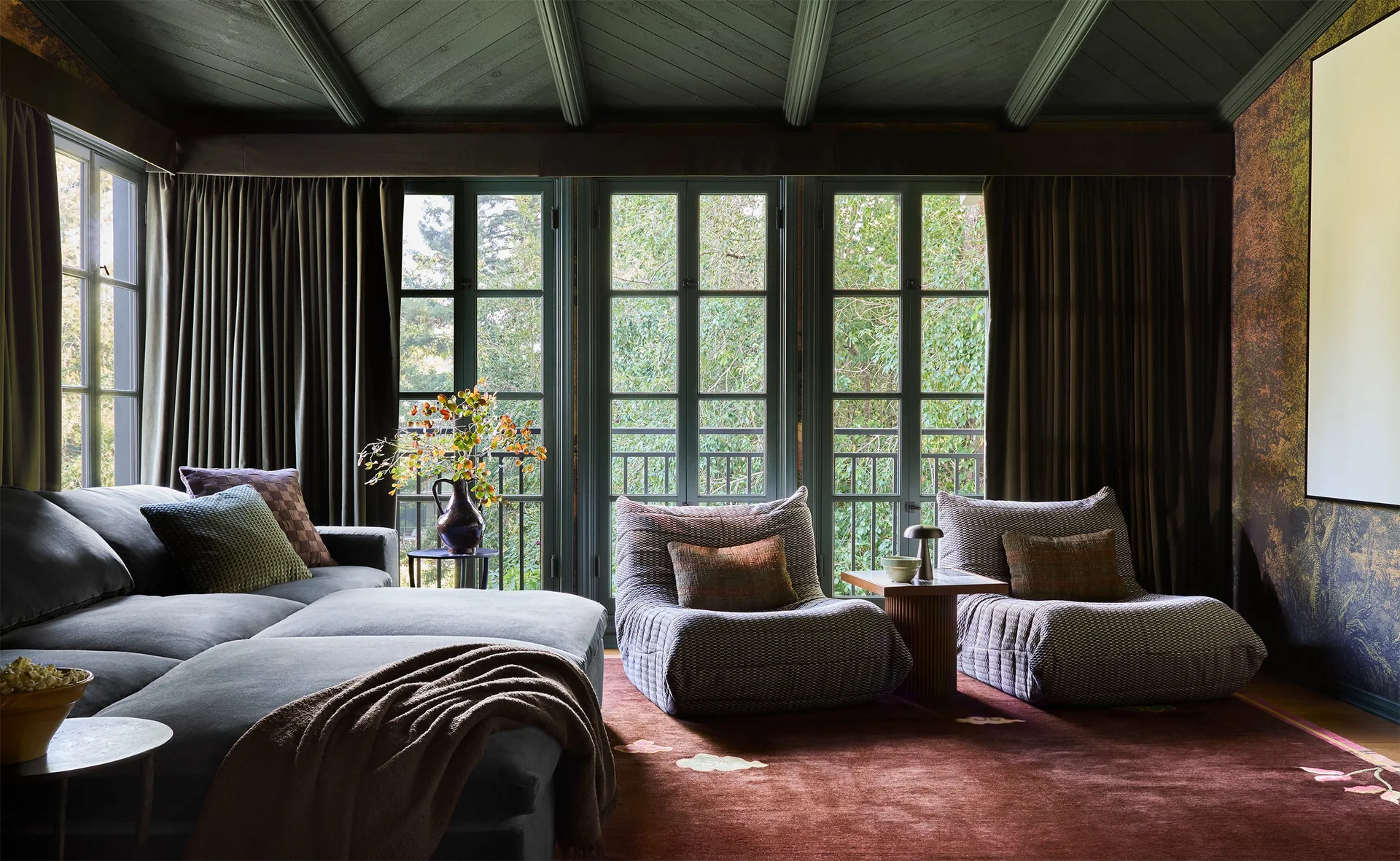 A moody living room features a dark sectional sofa, two patterned lounge chairs, a deep red rug, and dark green ceiling beams near large windows.