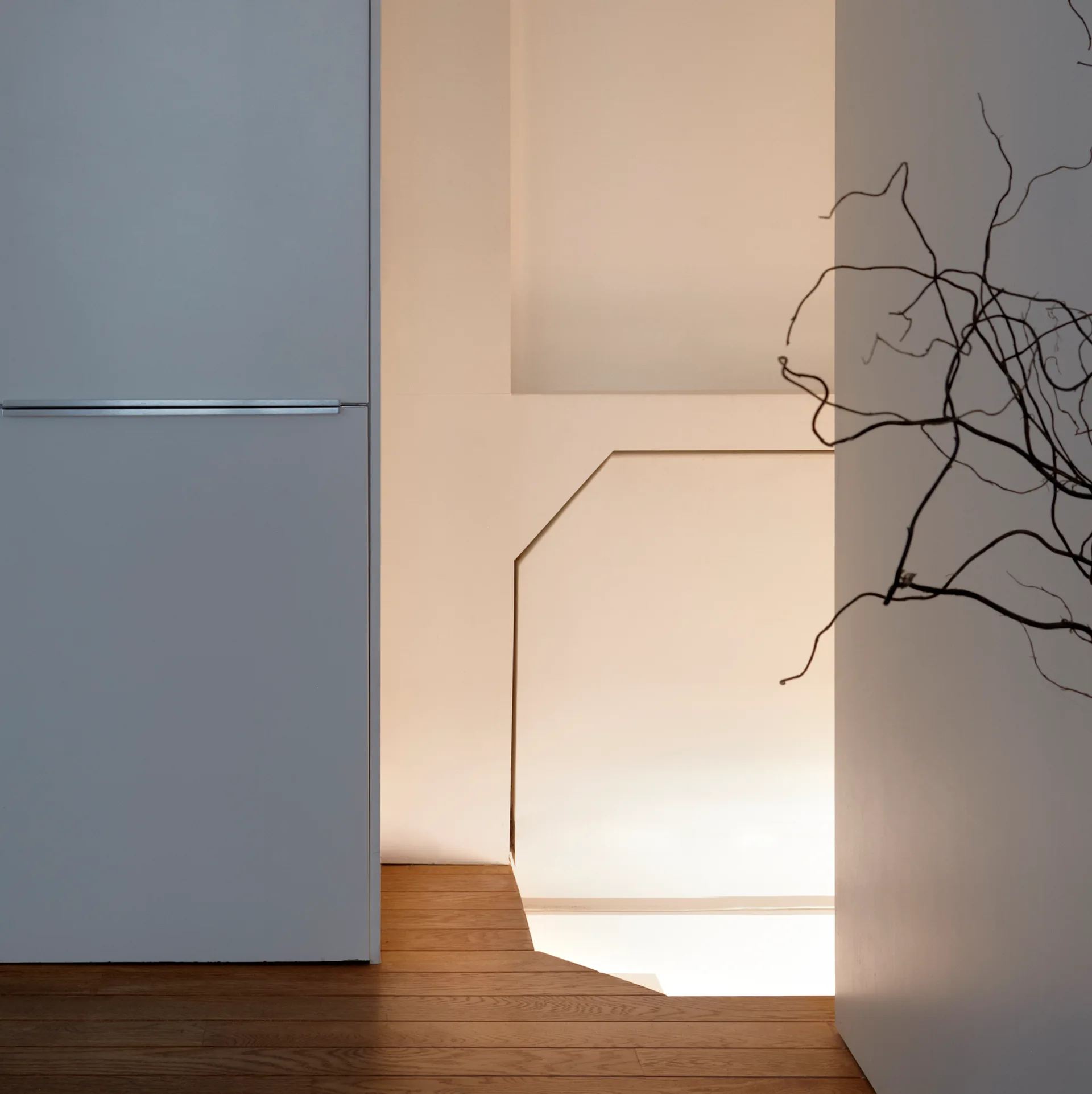 A minimalist hallway features wood flooring leading to an angled stairwell illuminated by warm recessed lighting next to a smooth grey panel.