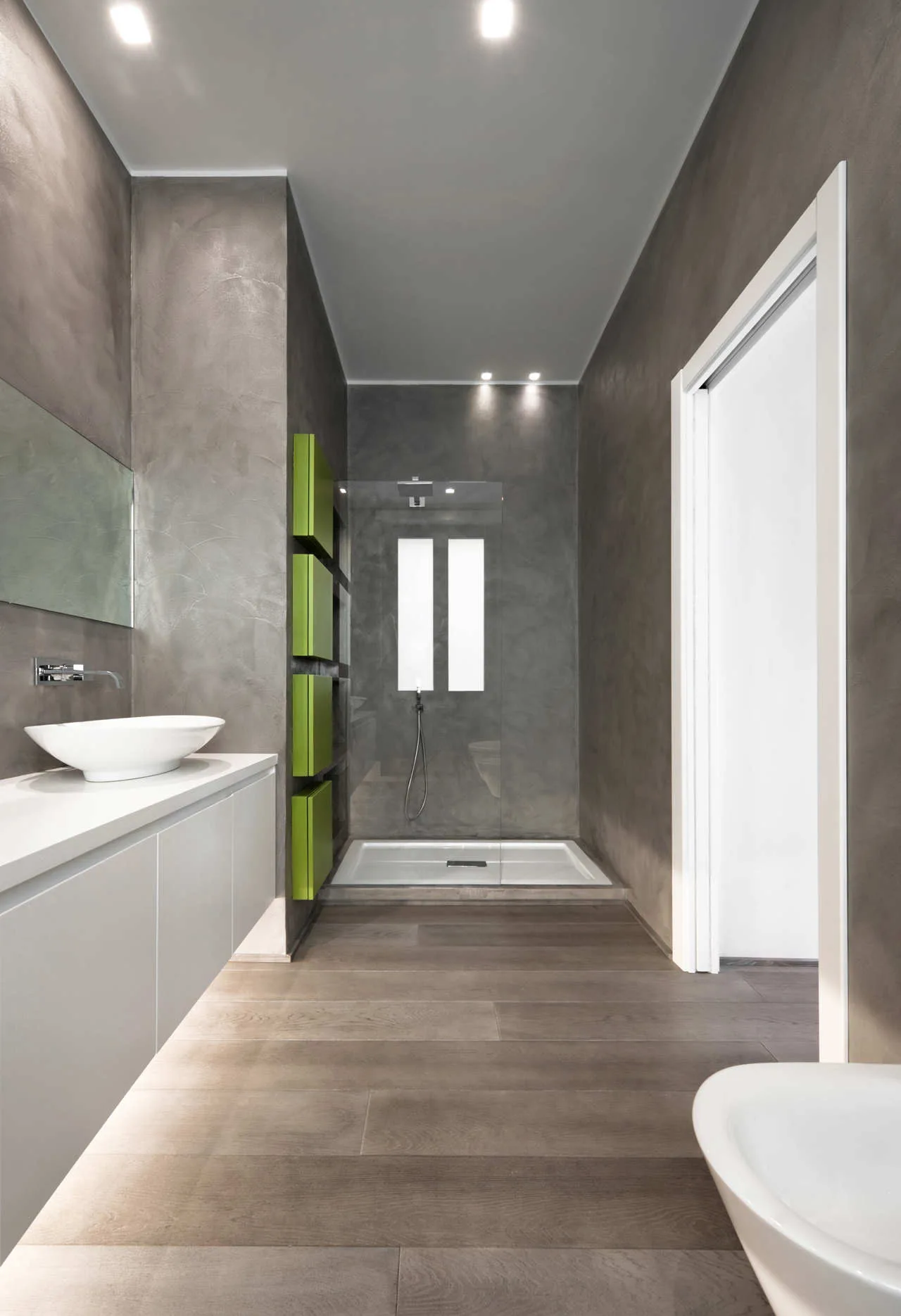 A modern bathroom with textured grey walls, a white floating vanity under a vessel sink, and recessed green shelves near a glass shower.