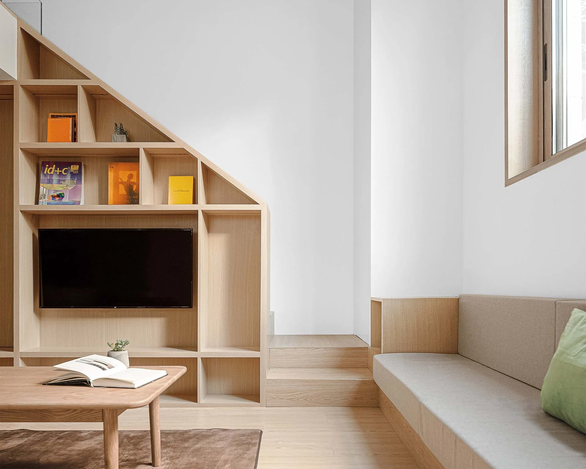 A minimalist living room shows a custom light wood under-stairs shelving unit with an integrated television, a built-in bench, and a coffee table.