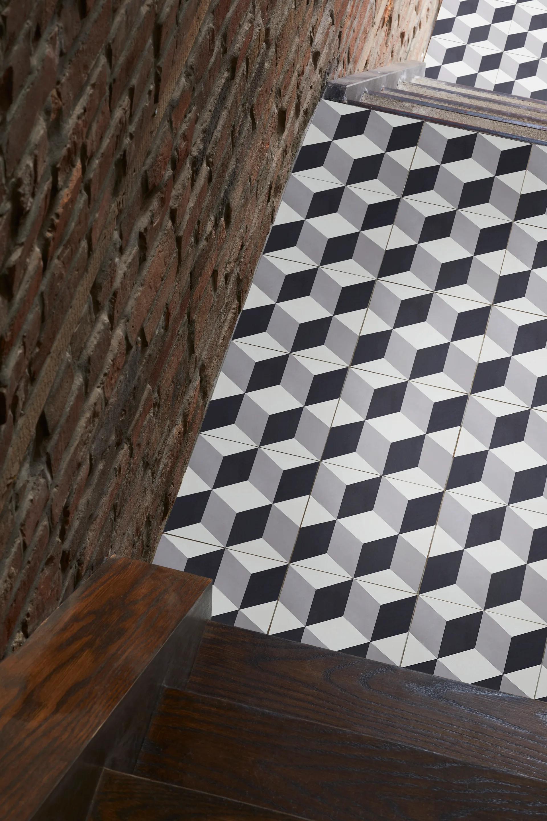 A staircase features dark wood treads and risers decorated with black, grey, and white geometric tile alongside an exposed red brick wall.