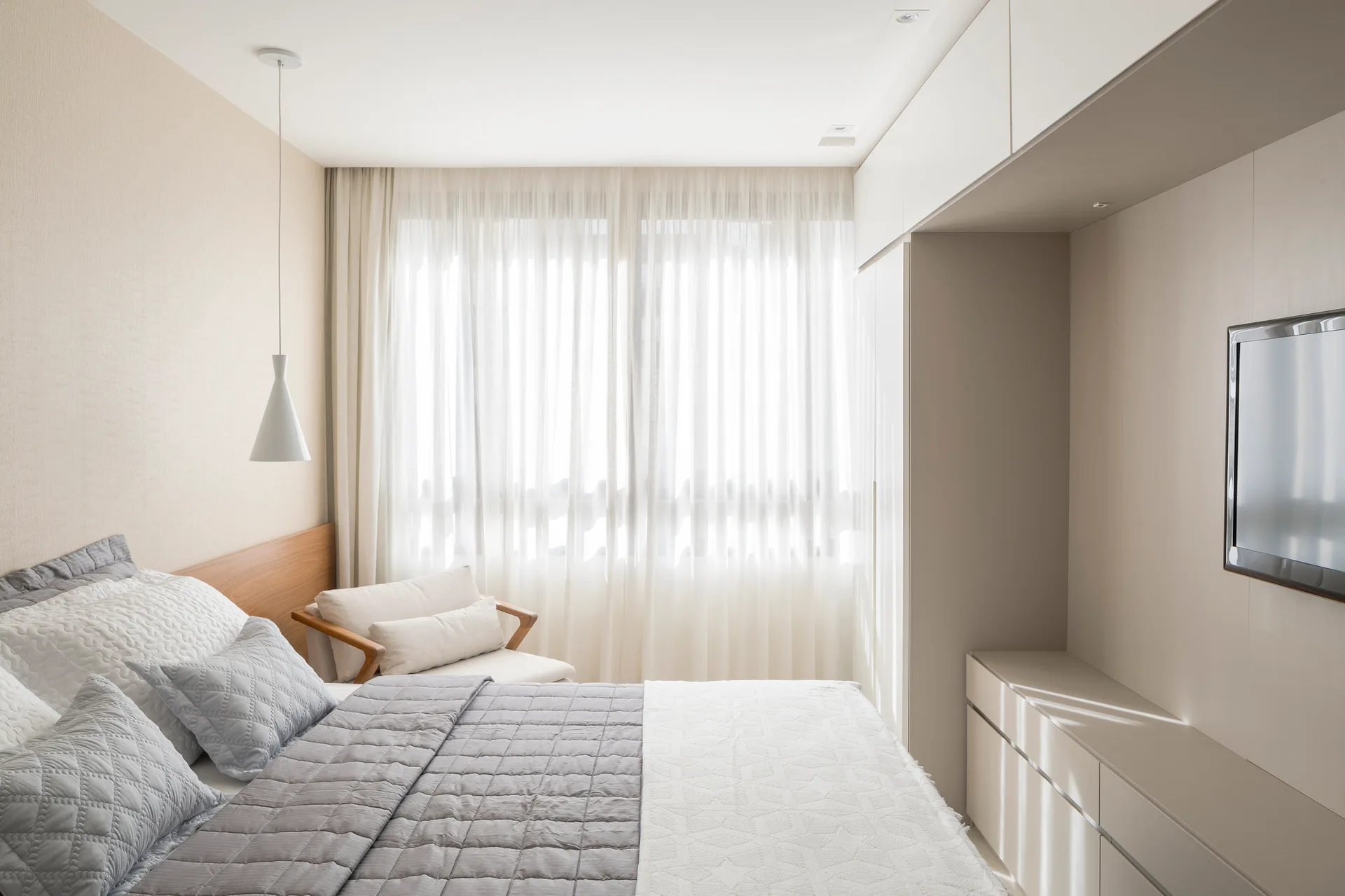 A modern bedroom featuring a bed with gray quilted covers, a light wood headboard, built-in beige wardrobes, and sheer white window curtains.