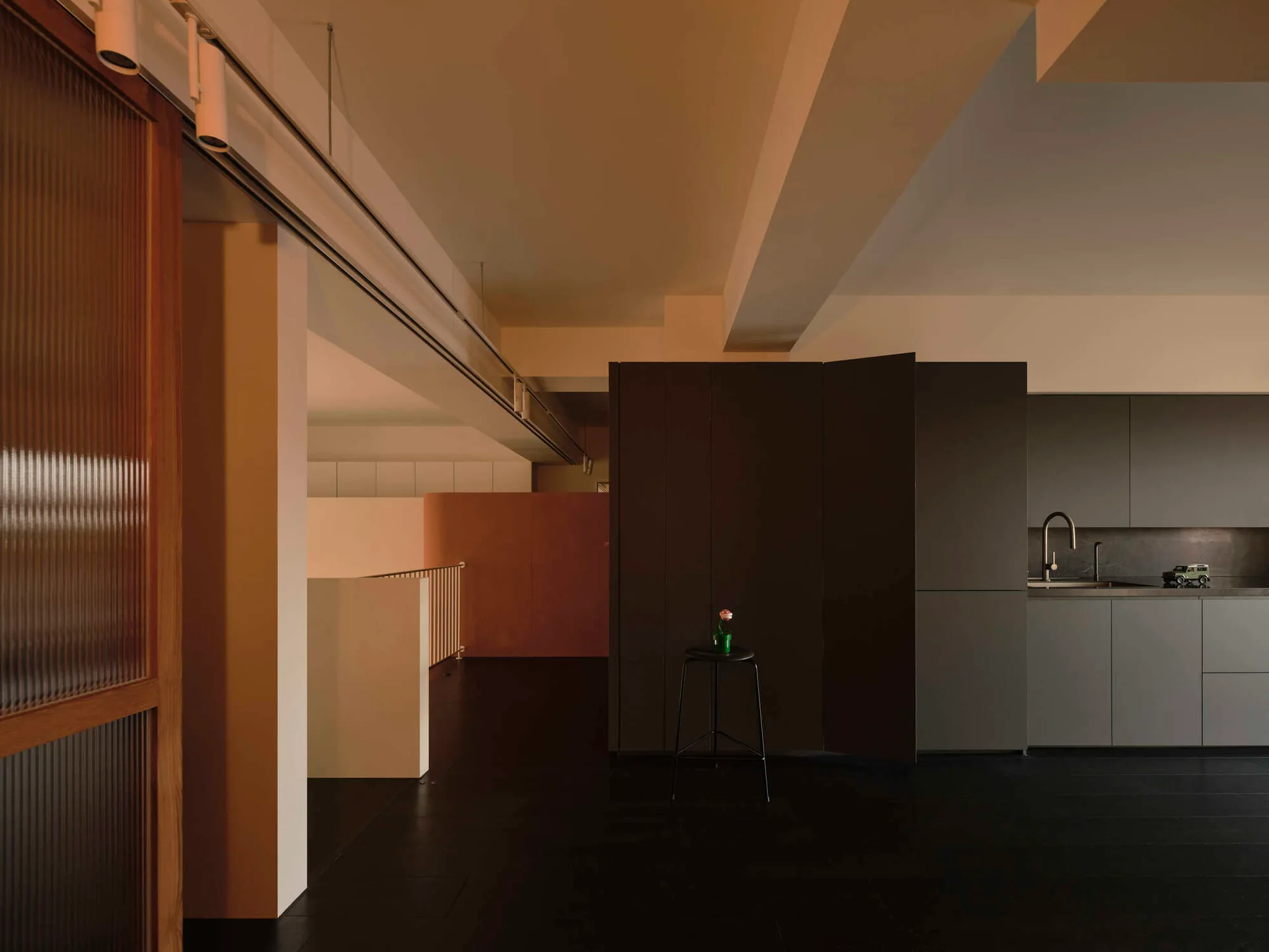 A minimalist kitchen features dark grey cabinetry and a sink alongside a black stool and a sliding door with ribbed glass panels under structural beams.