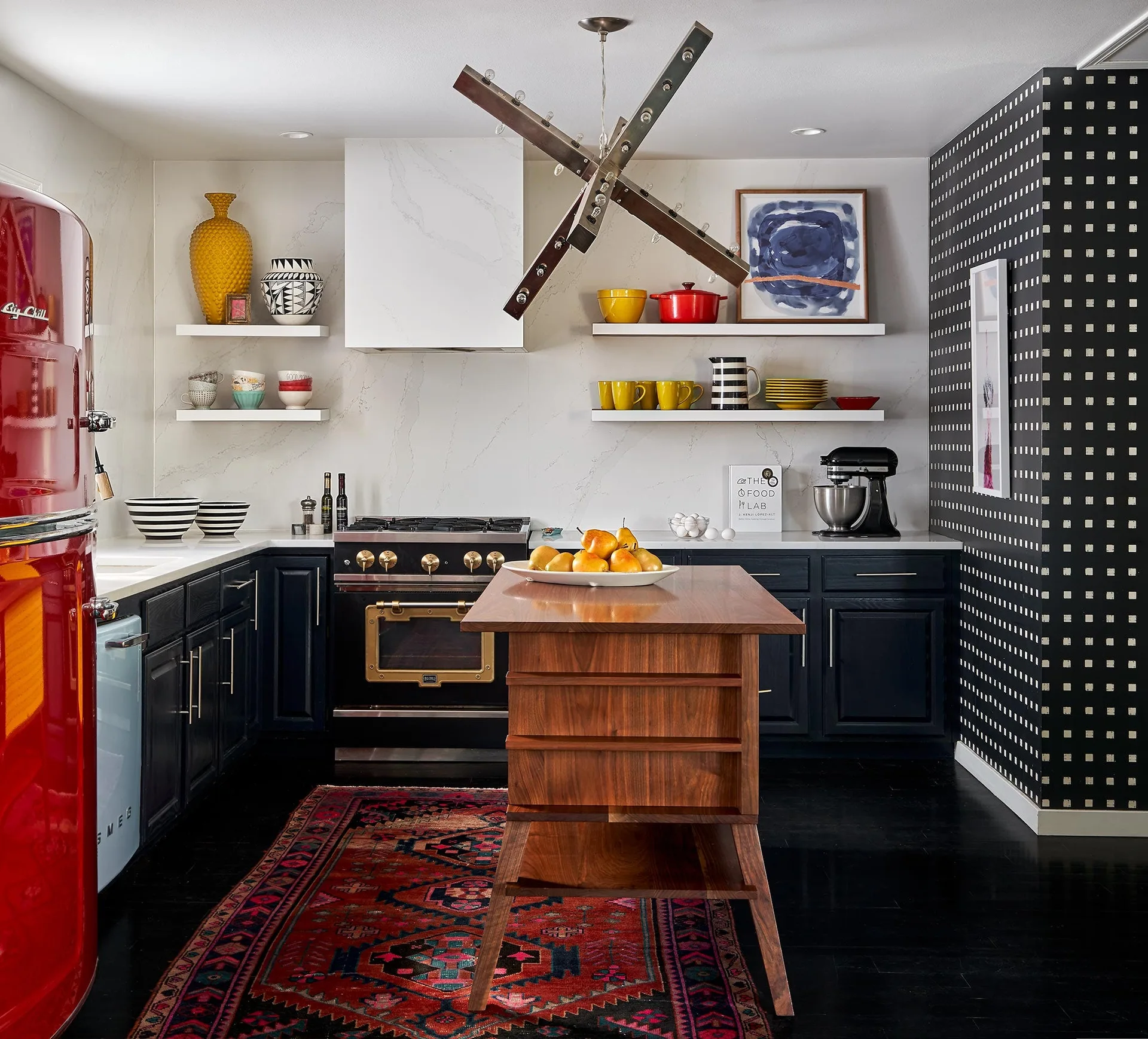 An eclectic kitchen features dark cabinetry, a wooden island, white floating shelves, a red vintage refrigerator, and a black patterned accent wall.