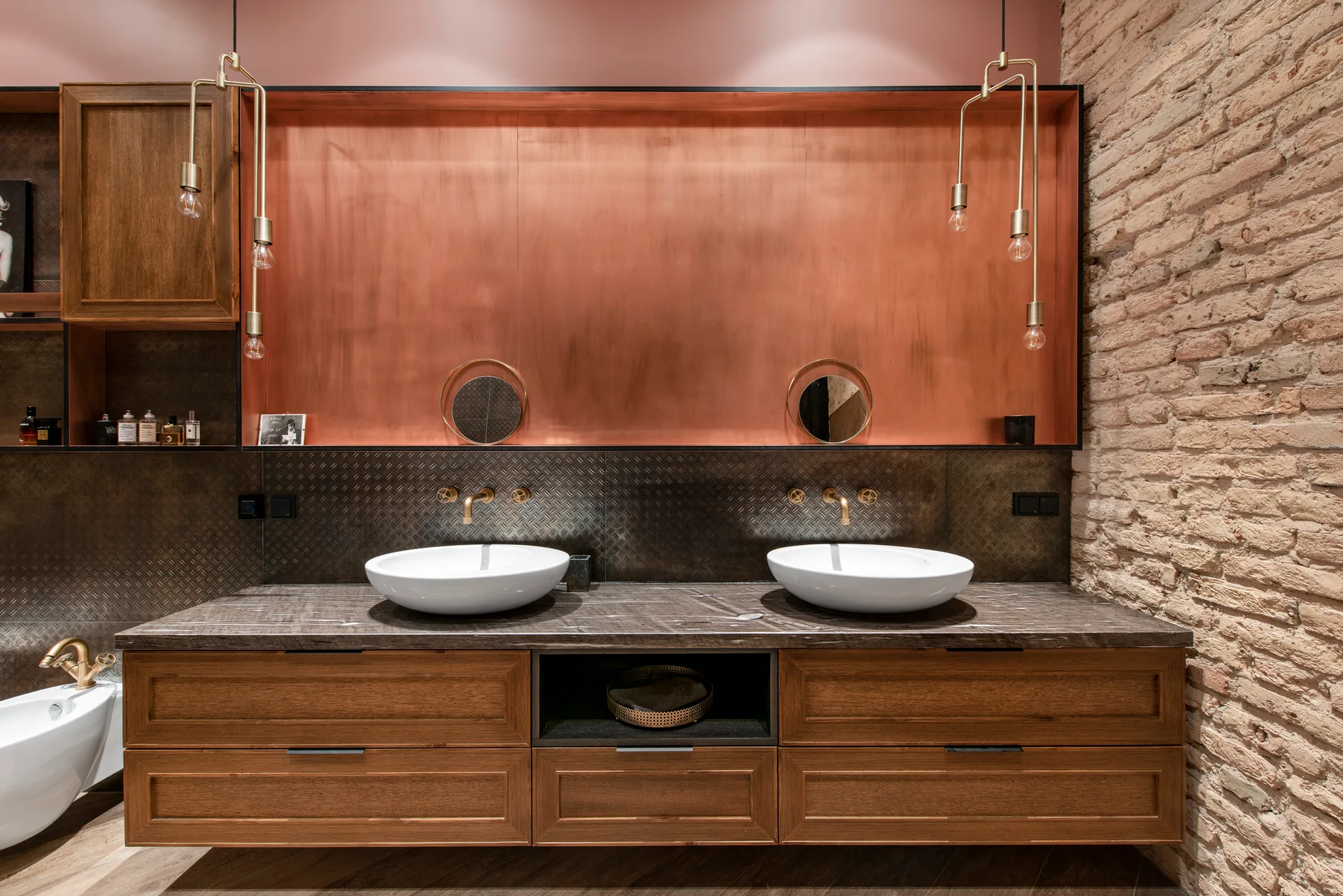 A bathroom vanity features two white vessel sinks, a copper metal backsplash, a wood floating cabinet, and an exposed brick accent wall.