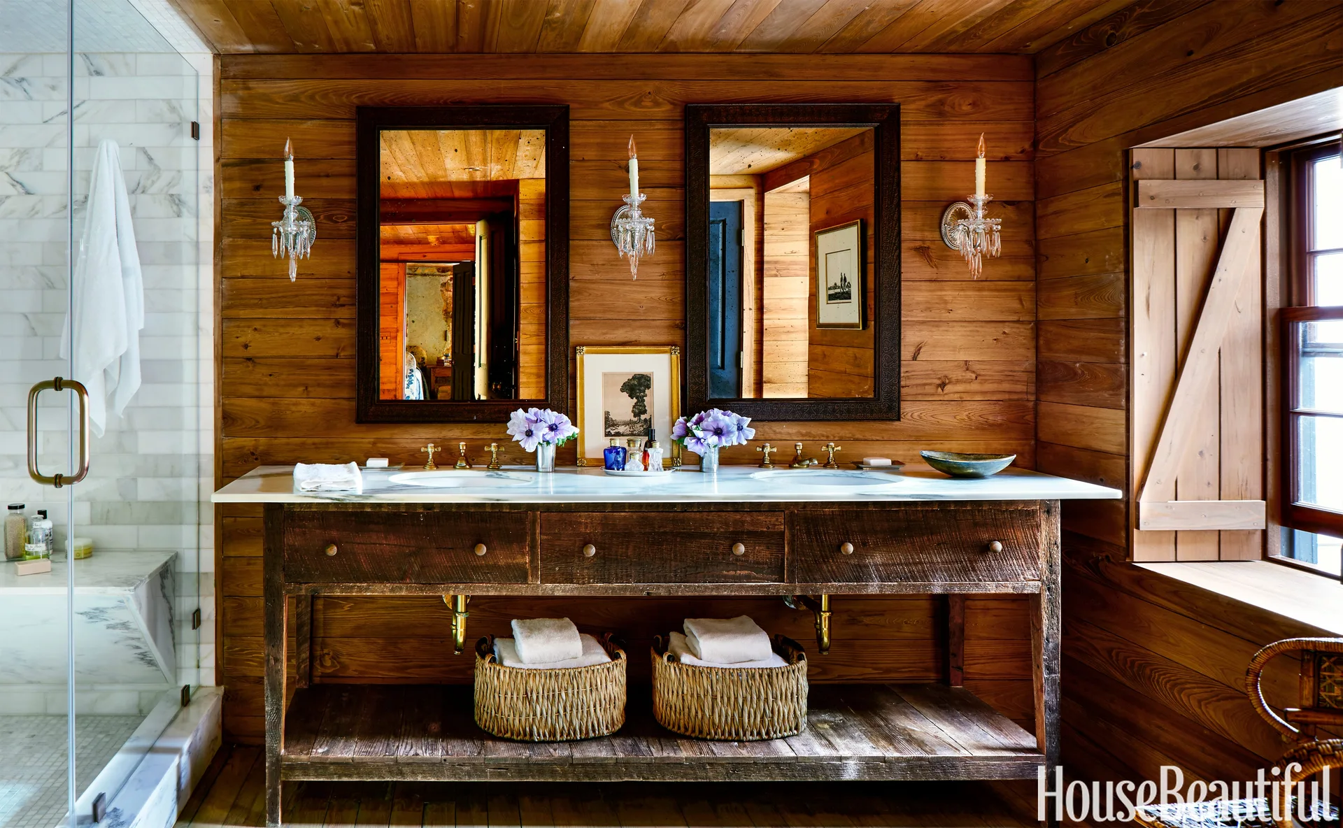 A rustic wood-paneled bathroom featuring a double sink vanity made of reclaimed wood, paired with a white marble countertop and crystal sconces.