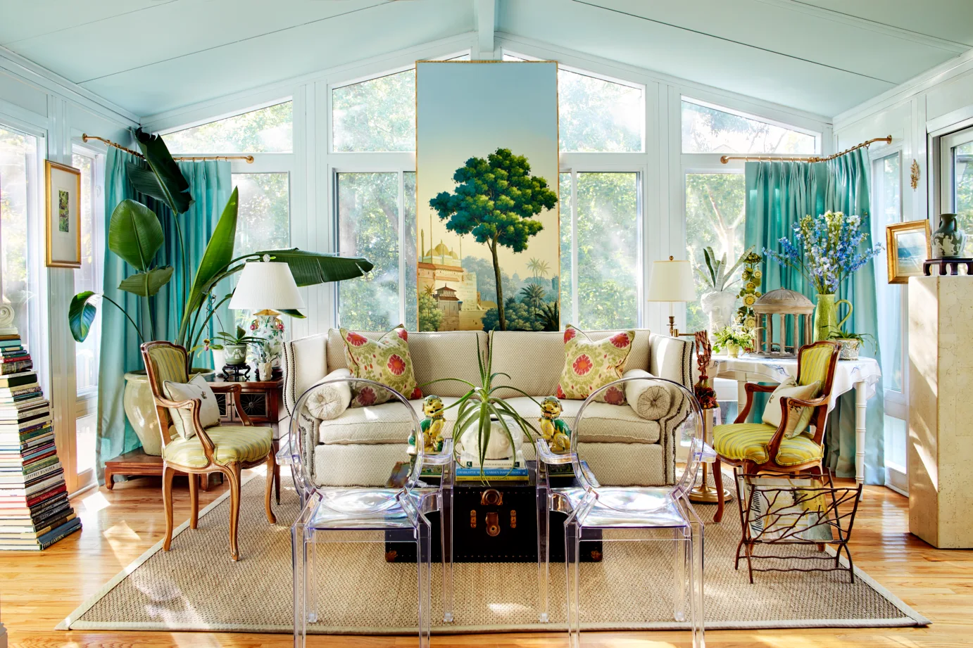 A sunroom with a cream sofa, teal curtains on large windows, a tall landscape painting, and two transparent acrylic chairs.