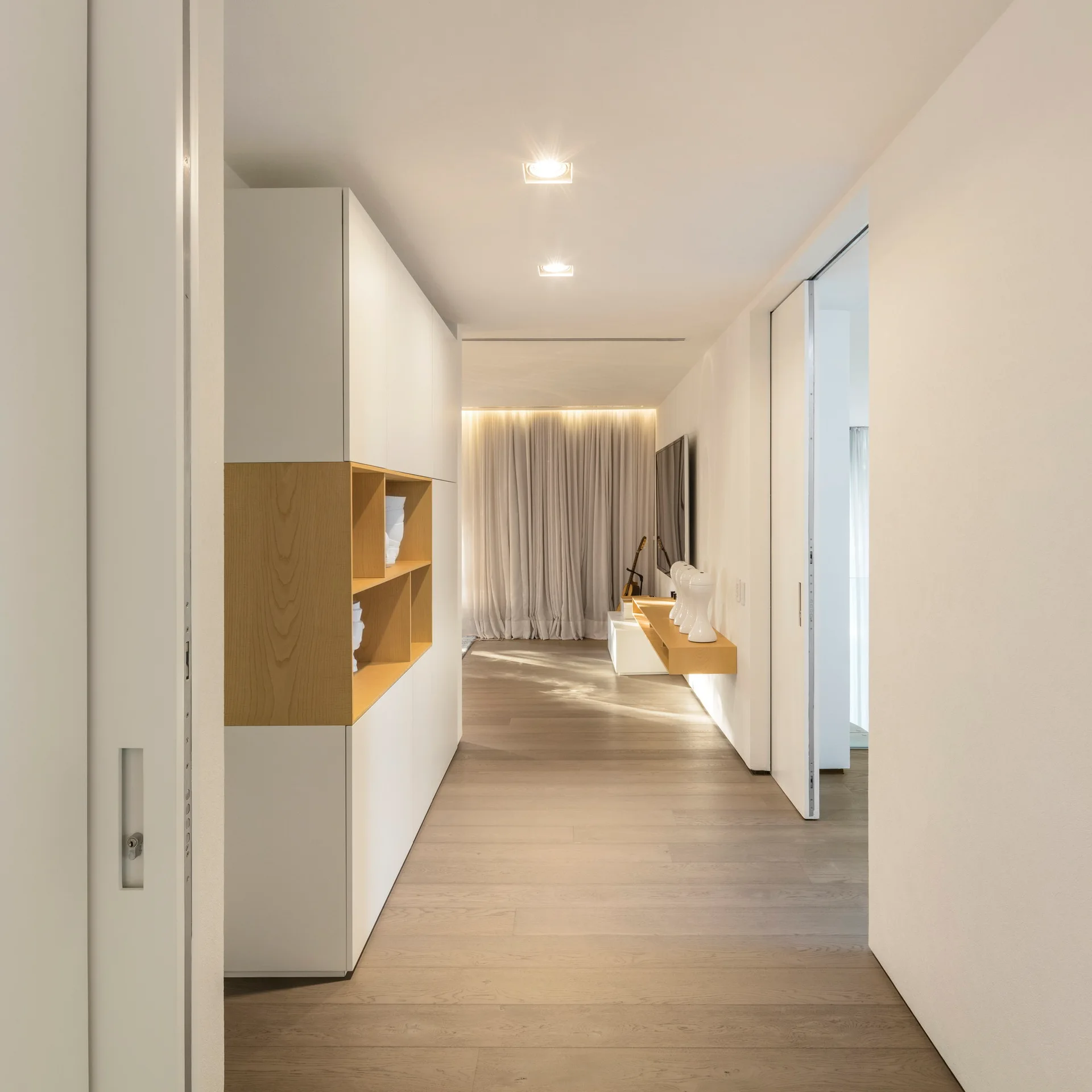 A minimalist hallway features built-in white storage cabinets with integrated wood shelving, light wood plank flooring, and soft recessed lighting.