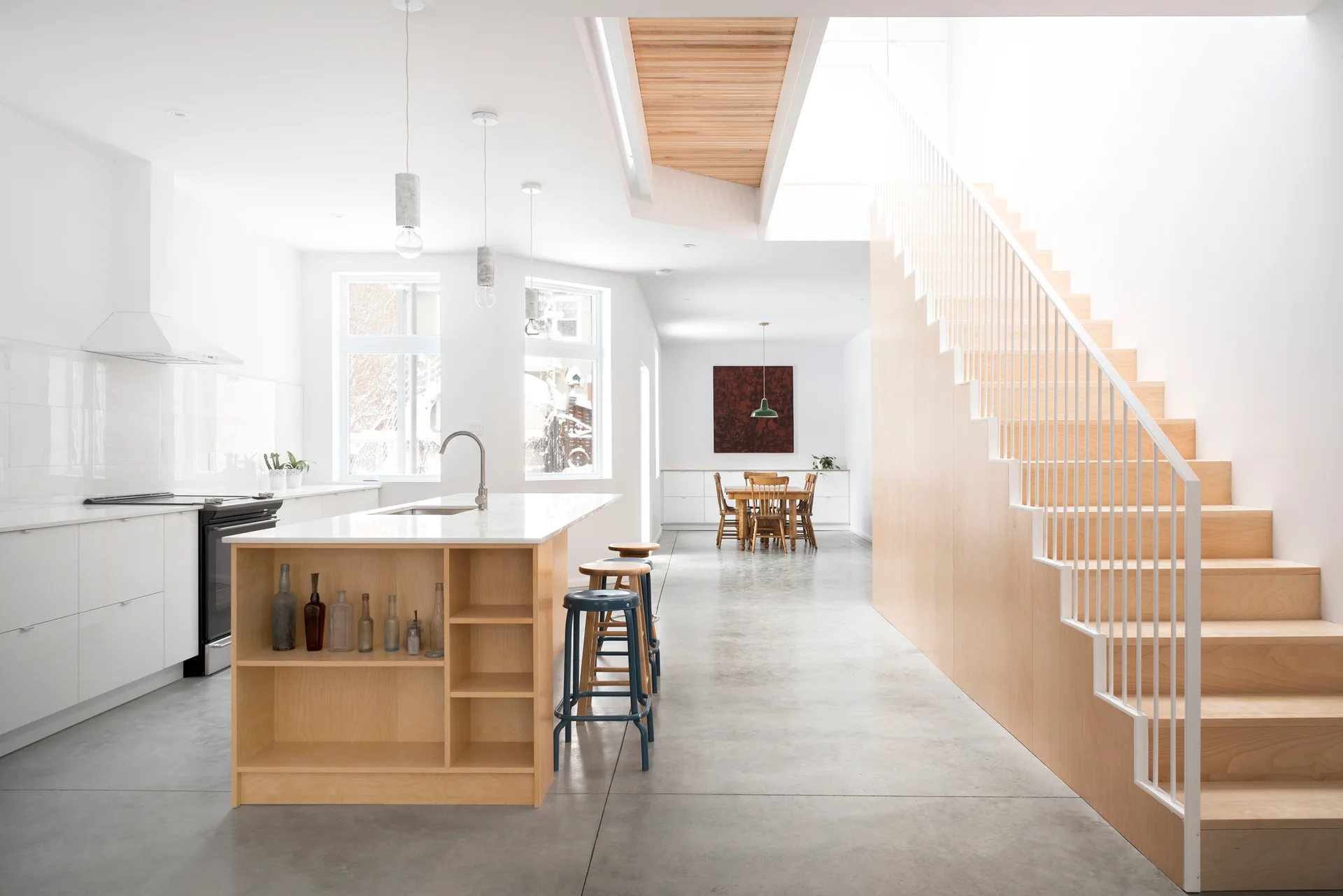 A minimalist open-concept kitchen features a light-toned plywood staircase, a matching wood kitchen island, white cabinets, and polished concrete flooring.