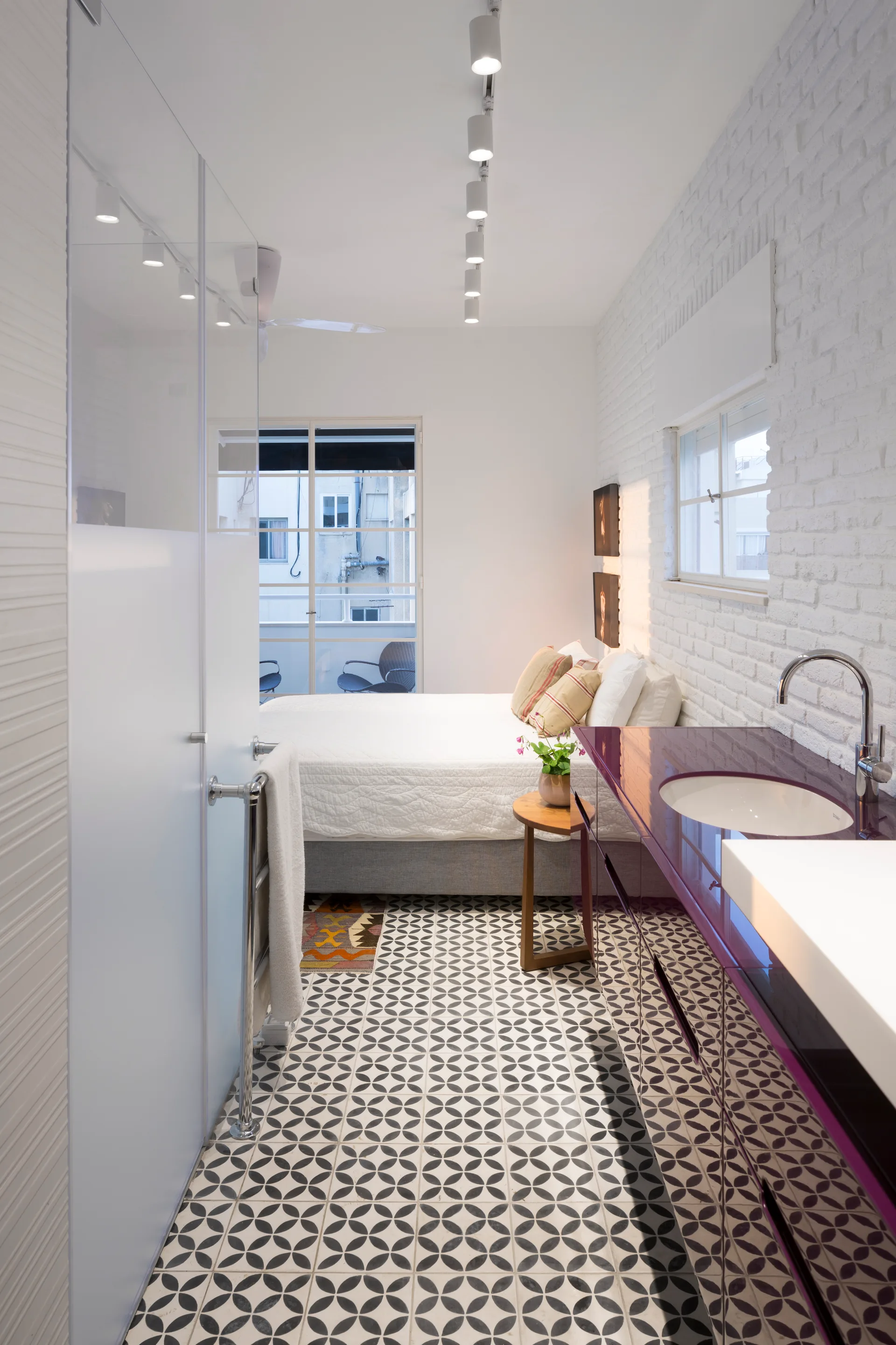 A long, narrow bathroom featuring a glossy purple vanity and patterned tile flooring that leads to an open bedroom.