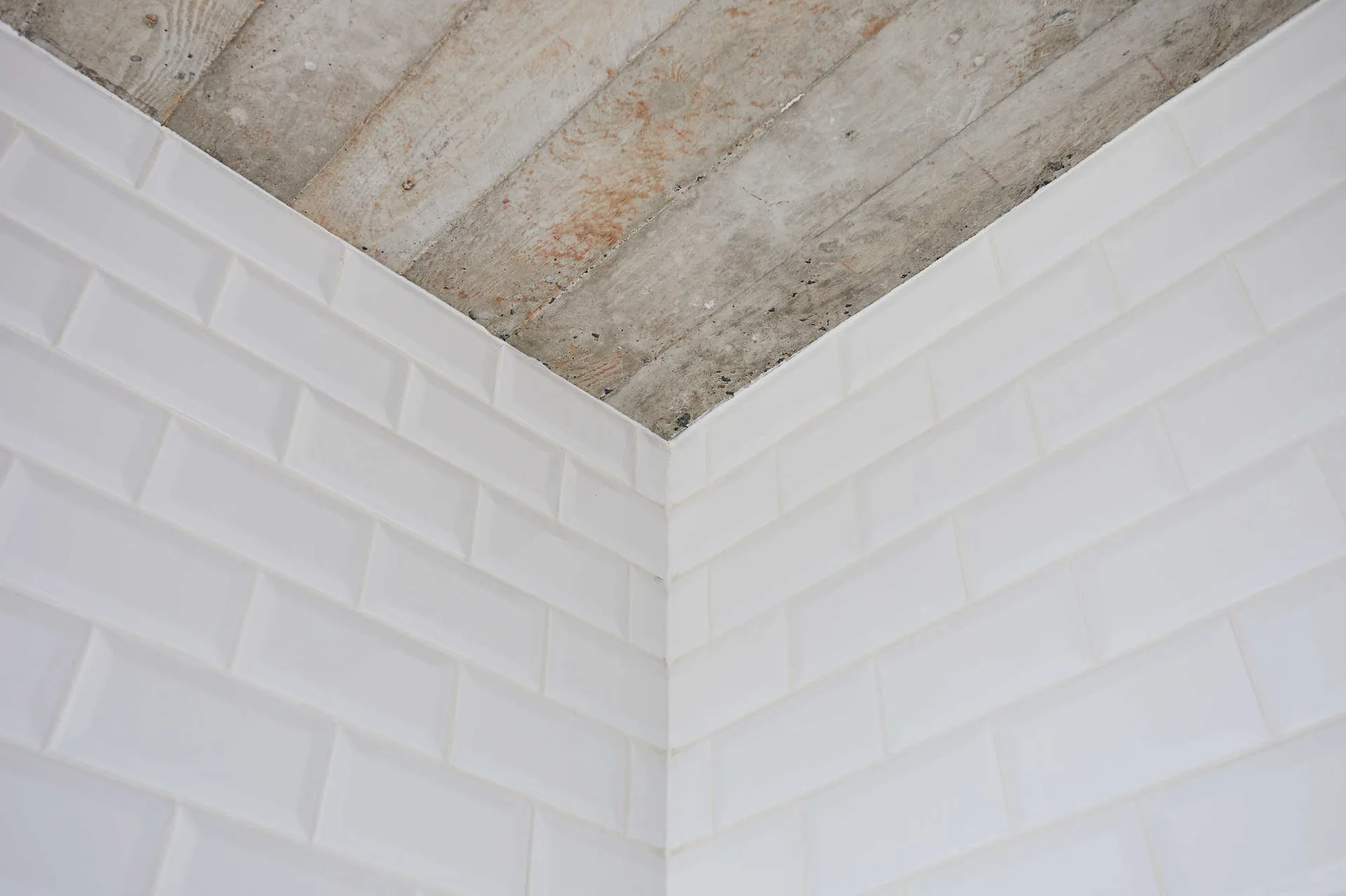 A close-up corner view showing white beveled subway tiles on the walls meeting a raw, board-formed concrete ceiling.