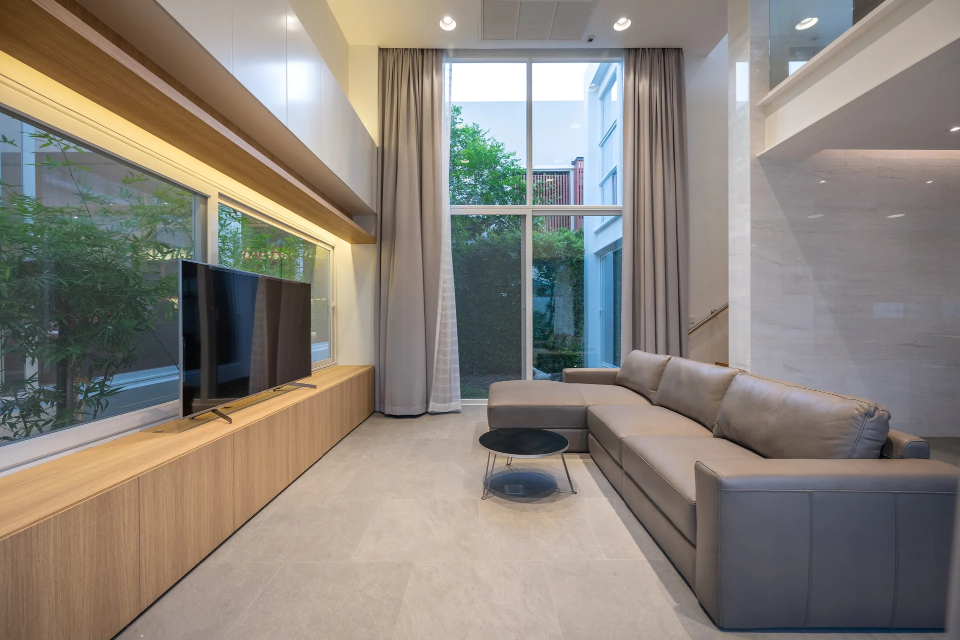 A modern living room features a grey leather sectional sofa, a long built-in wood console, and a tall floor-to-ceiling window with drapes.