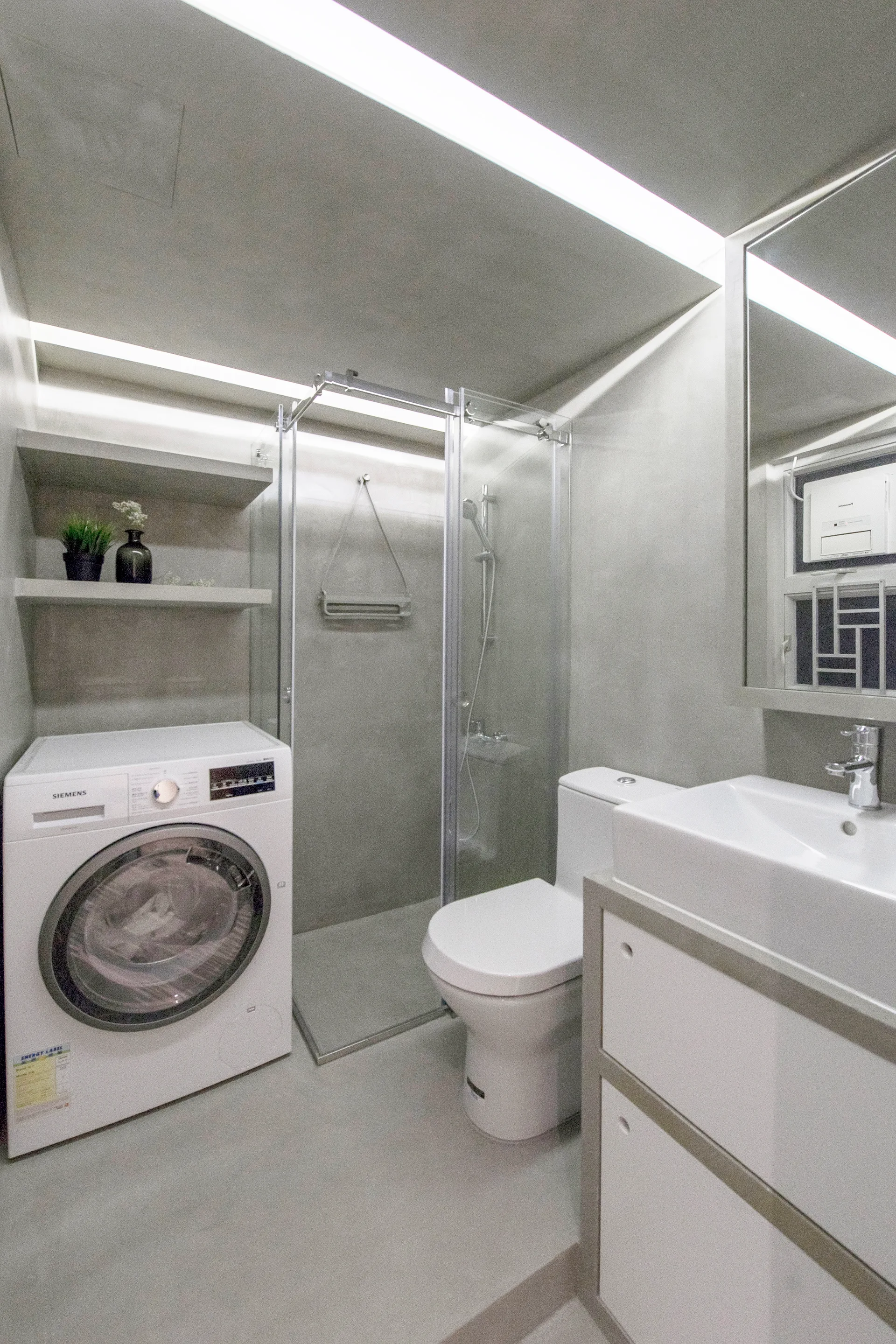 A modern grey concrete bathroom with a white washing machine, toilet, glass shower enclosure, and vanity with a rectangular mirror.