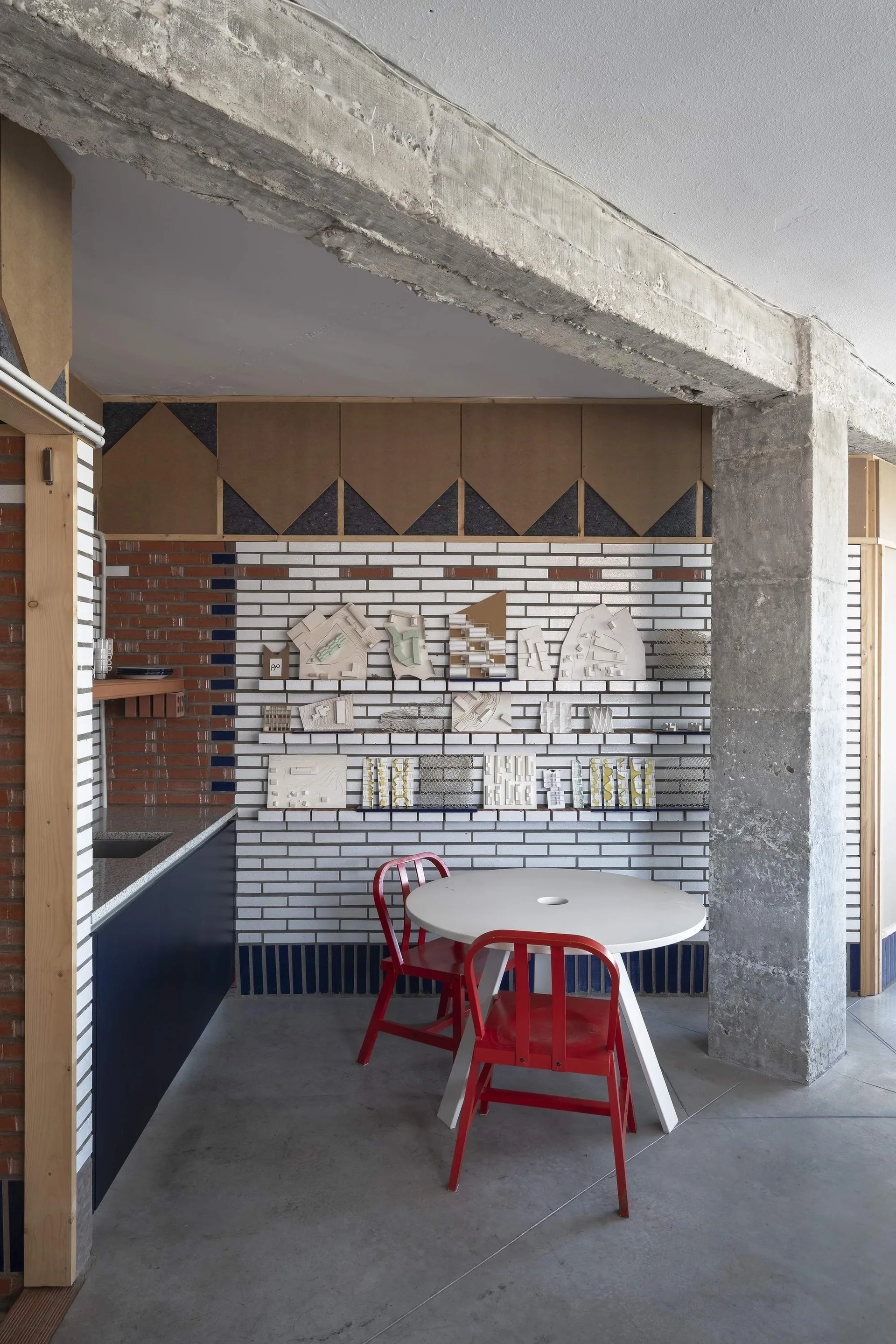 A circular white table and red metal chairs sit on a grey concrete floor next to an exposed concrete column and a white brick wall showing models.
