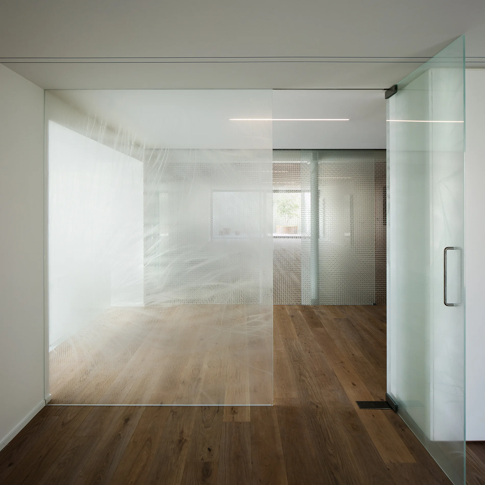 A minimalist interior space featuring patterned glass partition walls, an open glass door, and natural hardwood plank flooring.