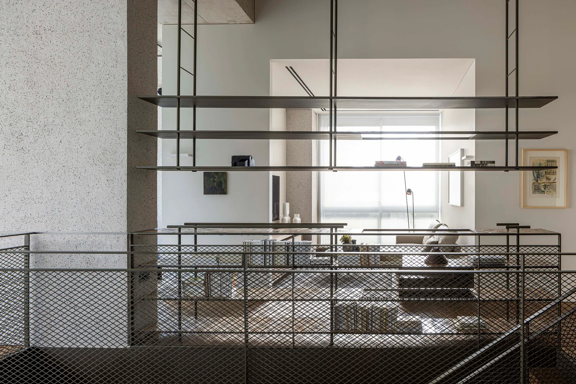 A view through a dark metal mesh railing toward a bright living room featuring suspended steel shelving, a sofa, and textured plaster walls.