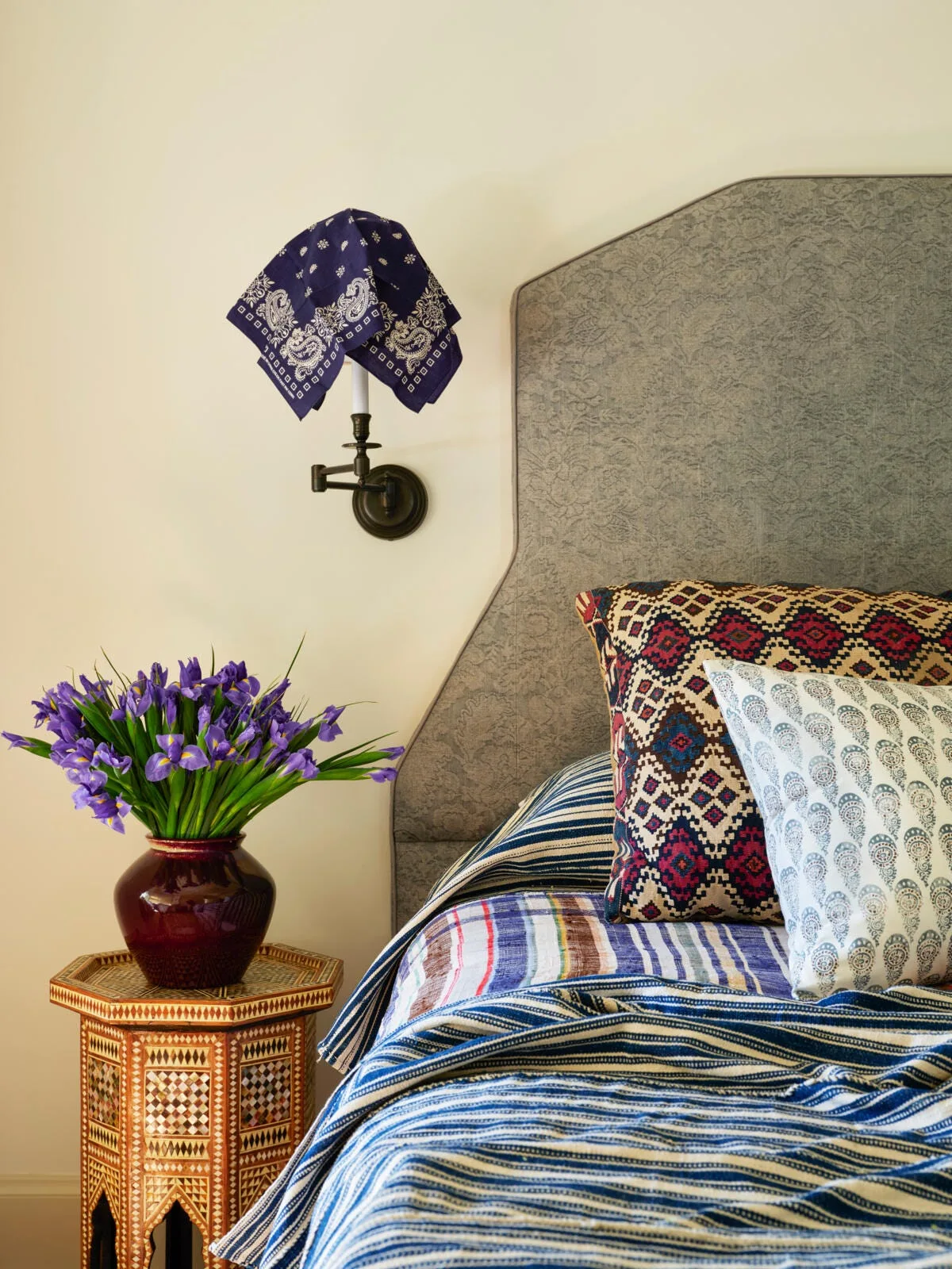 An eclectic bedroom corner features a gray upholstered headboard, blue striped bedding, an inlay side table with purple irises, and a wall sconce.