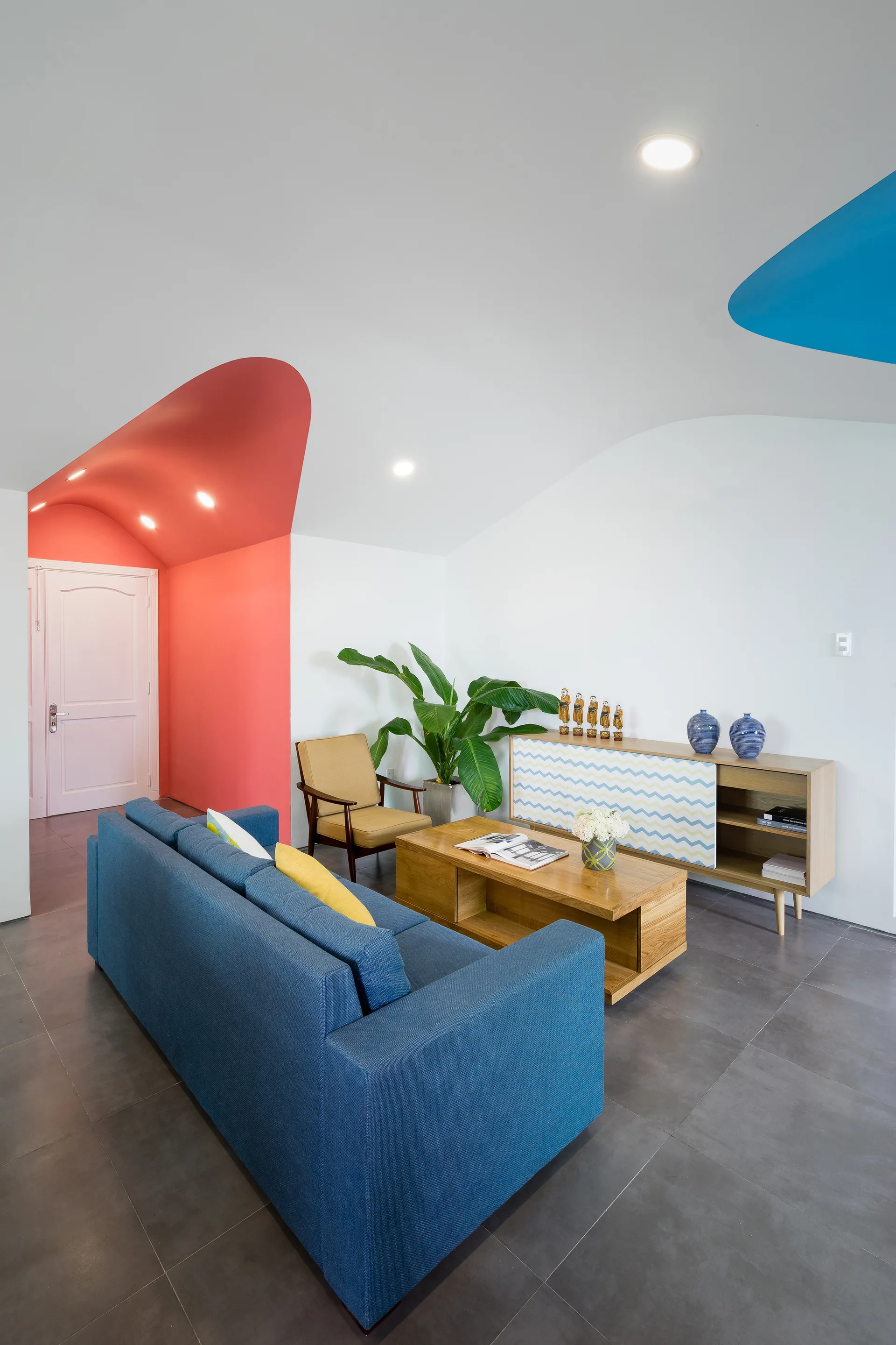 A modern living room features a blue sofa, a wood coffee table, and a curved white ceiling painted with bold red and blue accents.