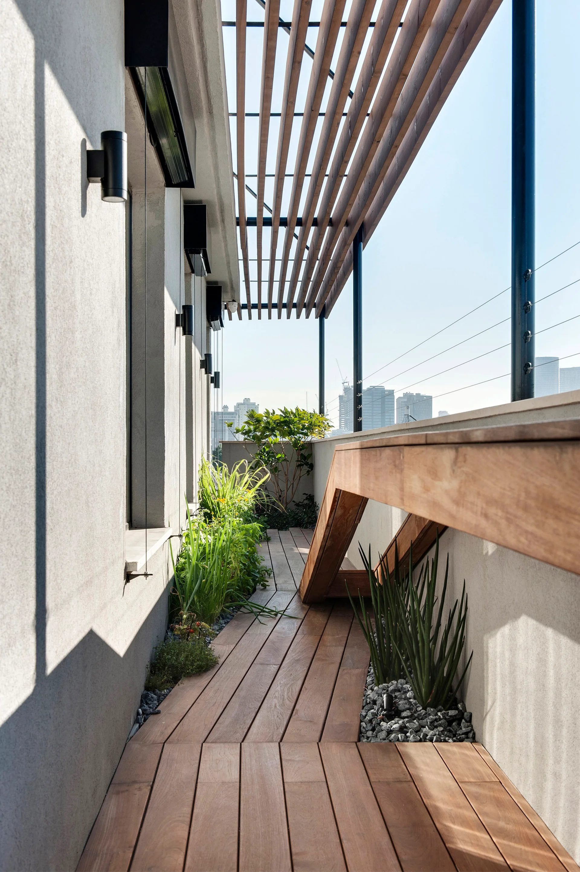 A narrow outdoor balcony features a wooden plank deck, an overhead slatted pergola, gray concrete walls, and small integrated planters with green plants.