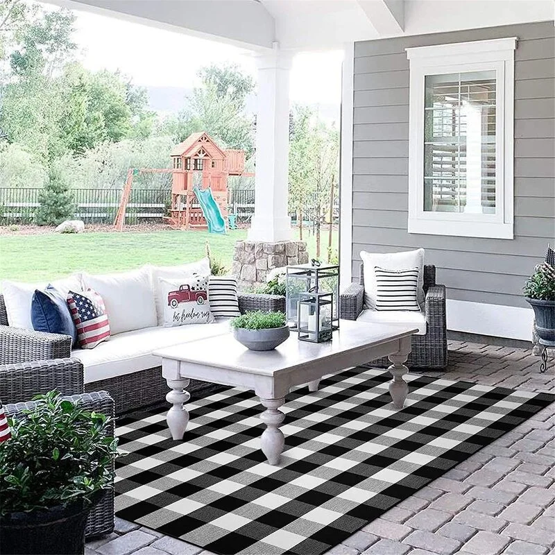 A covered patio features a grey wicker sectional sofa, a white wood coffee table, and a large black and white buffalo plaid rug on stone pavers.