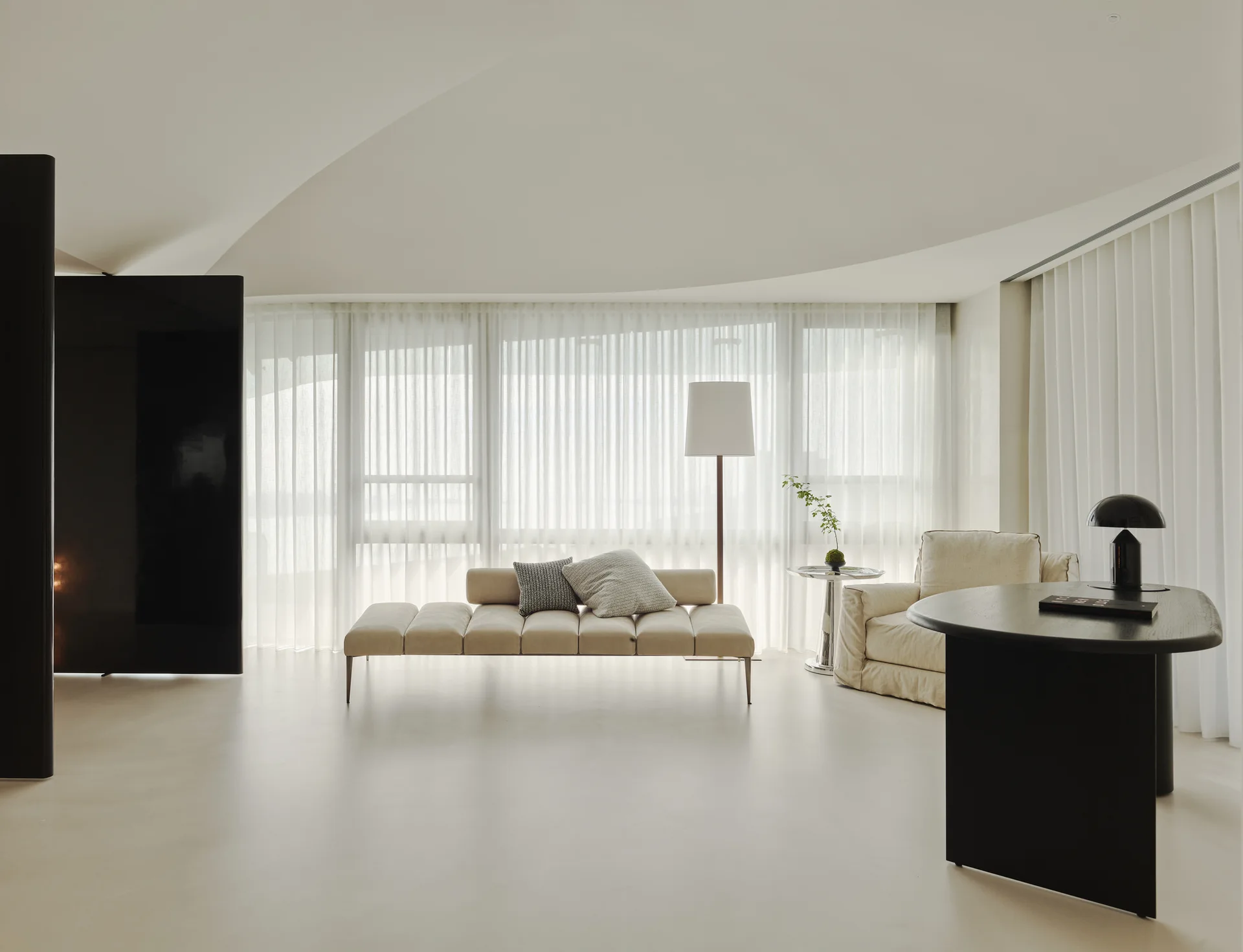 A minimalist living space featuring a cream tufted daybed, a curved black desk, and large windows with sheer white drapes under a curved ceiling.