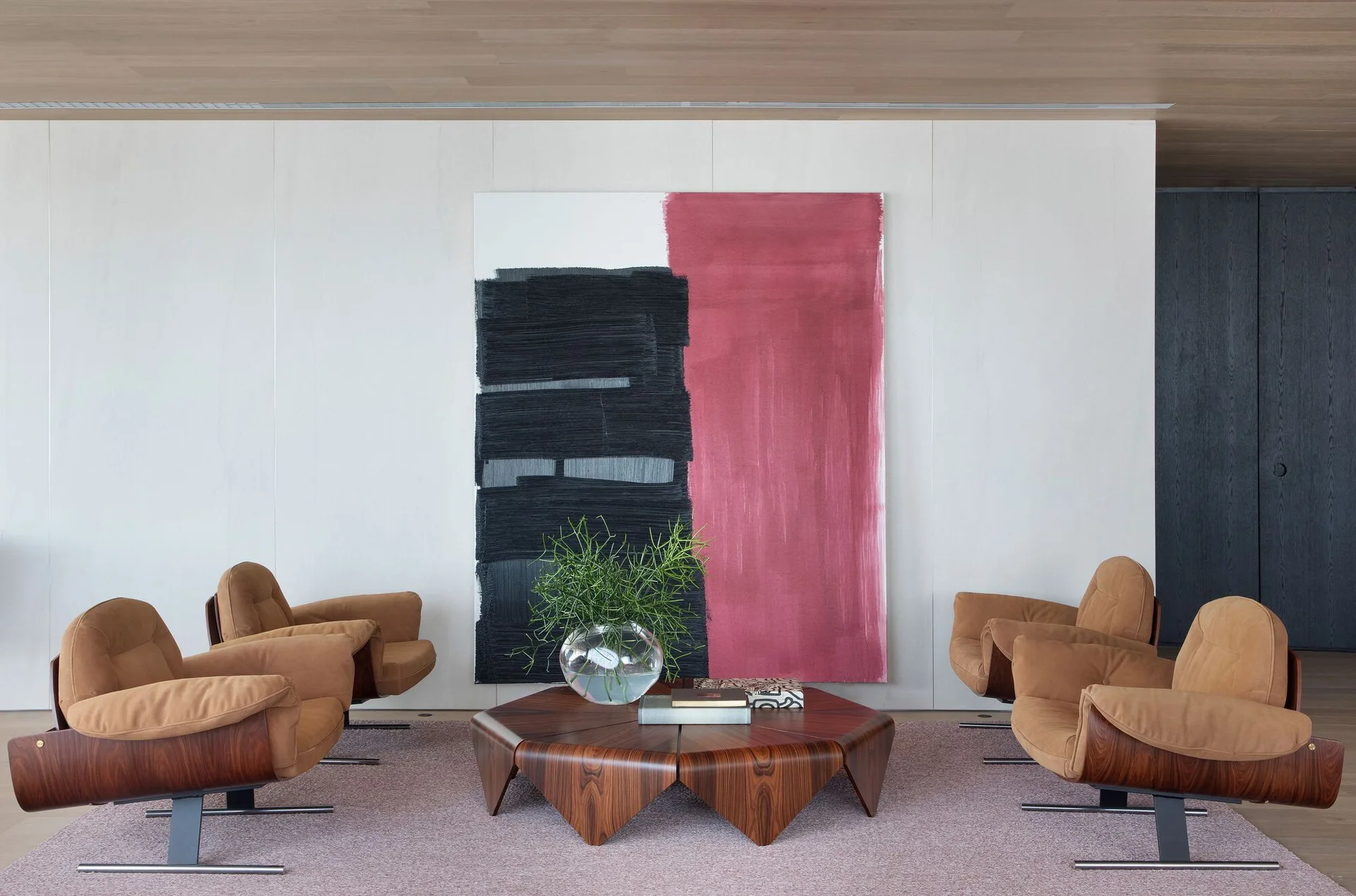 Four tan armchairs surround a geometric wood coffee table in front of a large red and black abstract painting.