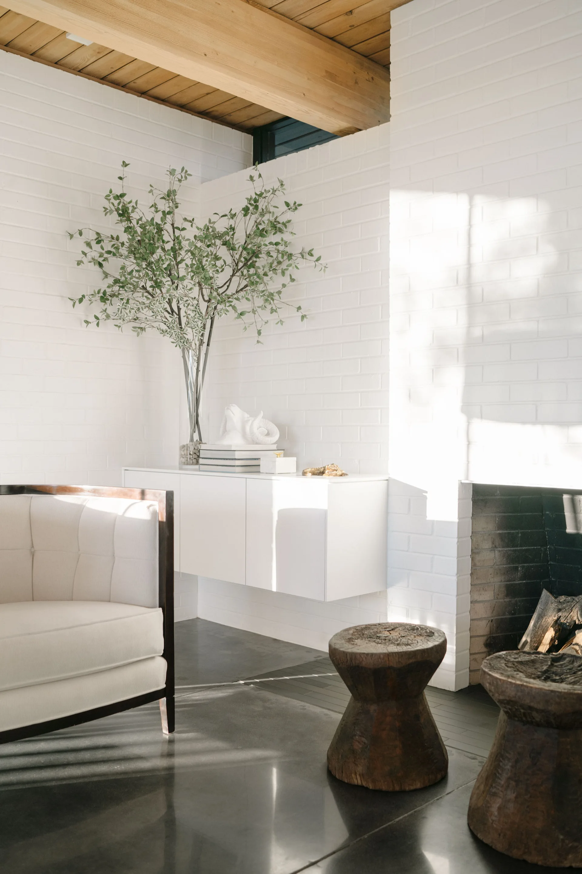 A modern living room features a white painted brick wall with a fireplace, a floating console, a cream armchair, and carved wooden stools.