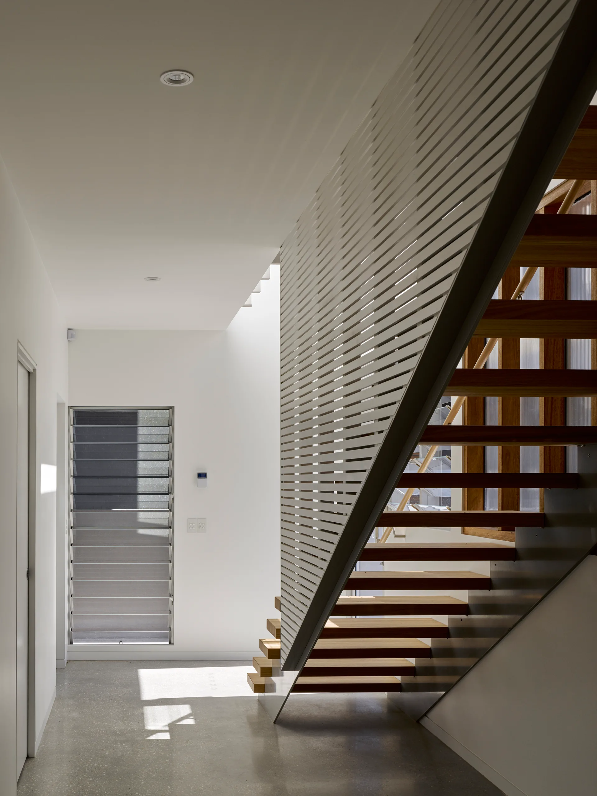 A modern open-tread wooden staircase is framed by a white horizontal slat screen and sits beside a tall louvered window on a polished concrete floor.