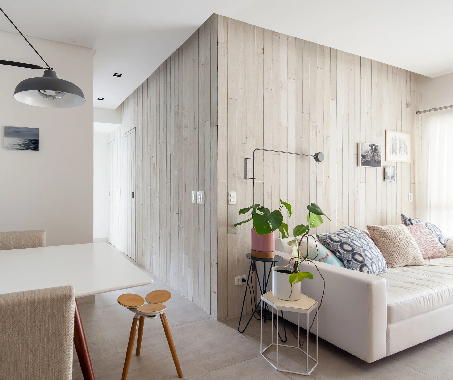 A modern living space features a white sofa, a vertical light wood plank accent wall, and nested plant stands beside a white dining table.