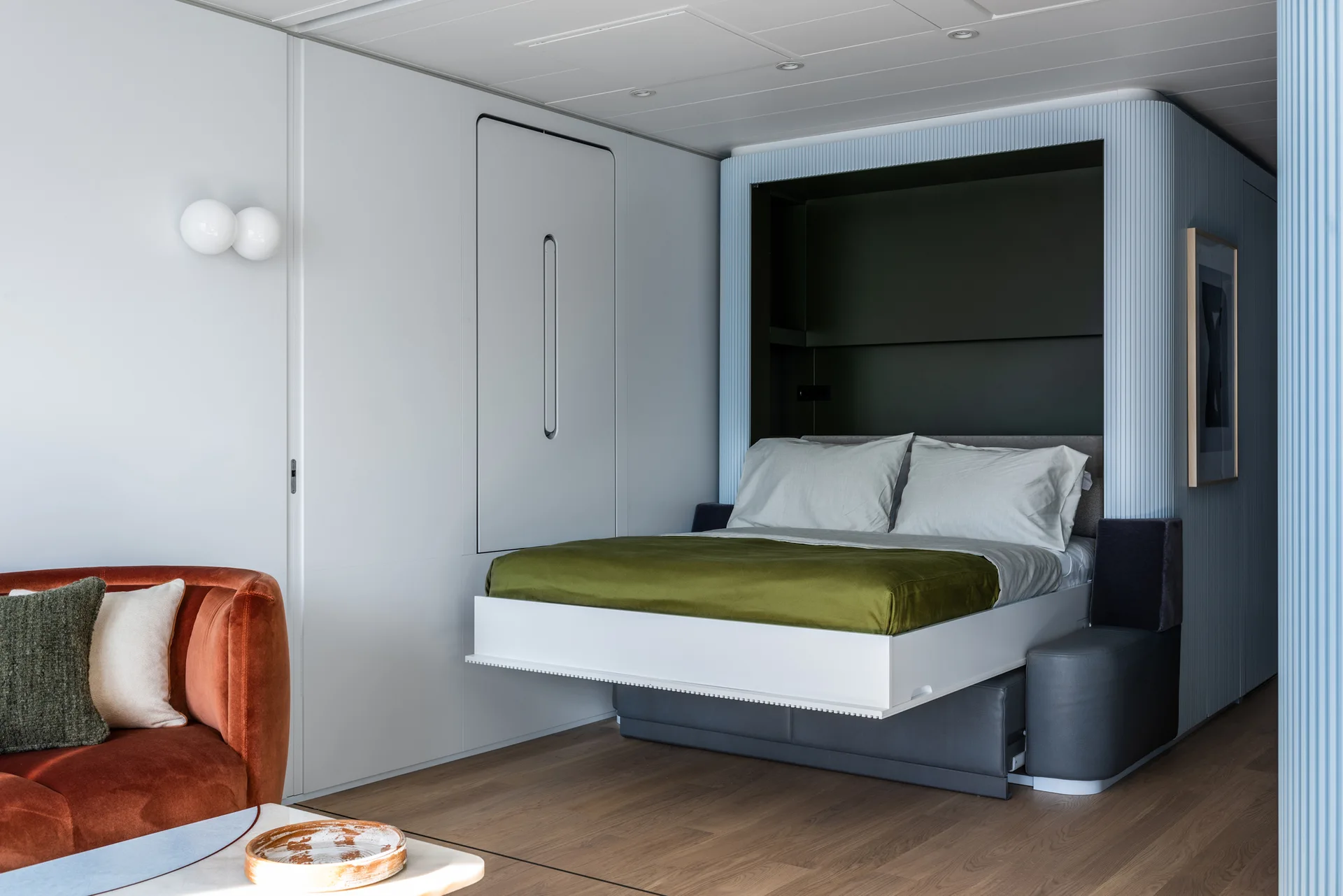 A modern bedroom features an integrated Murphy bed lowering over a grey base beside light blue fluted wall paneling and a rust velvet sofa.