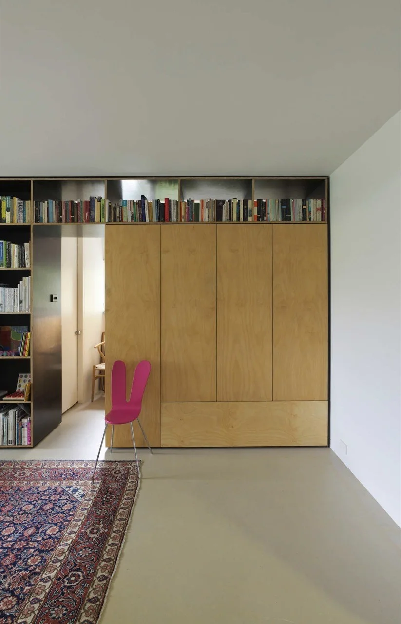 Minimalist room featuring built-in plywood cabinets, a top-running open bookshelf, an integrated doorway, and a bright pink accent chair.