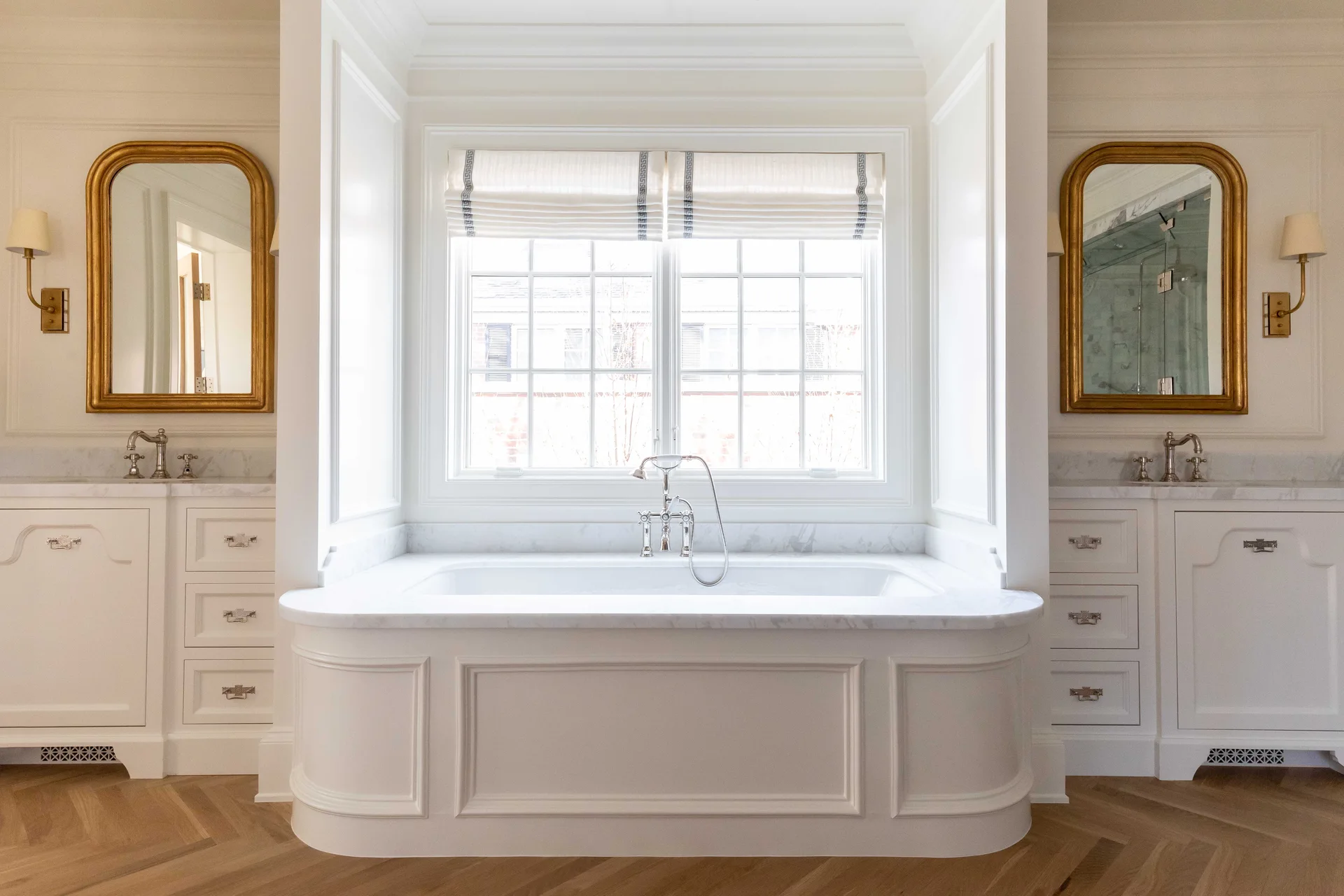Symmetrical white bathroom with a central built-in tub under a window, flanked by two vanities with arched gold mirrors on herringbone floors.