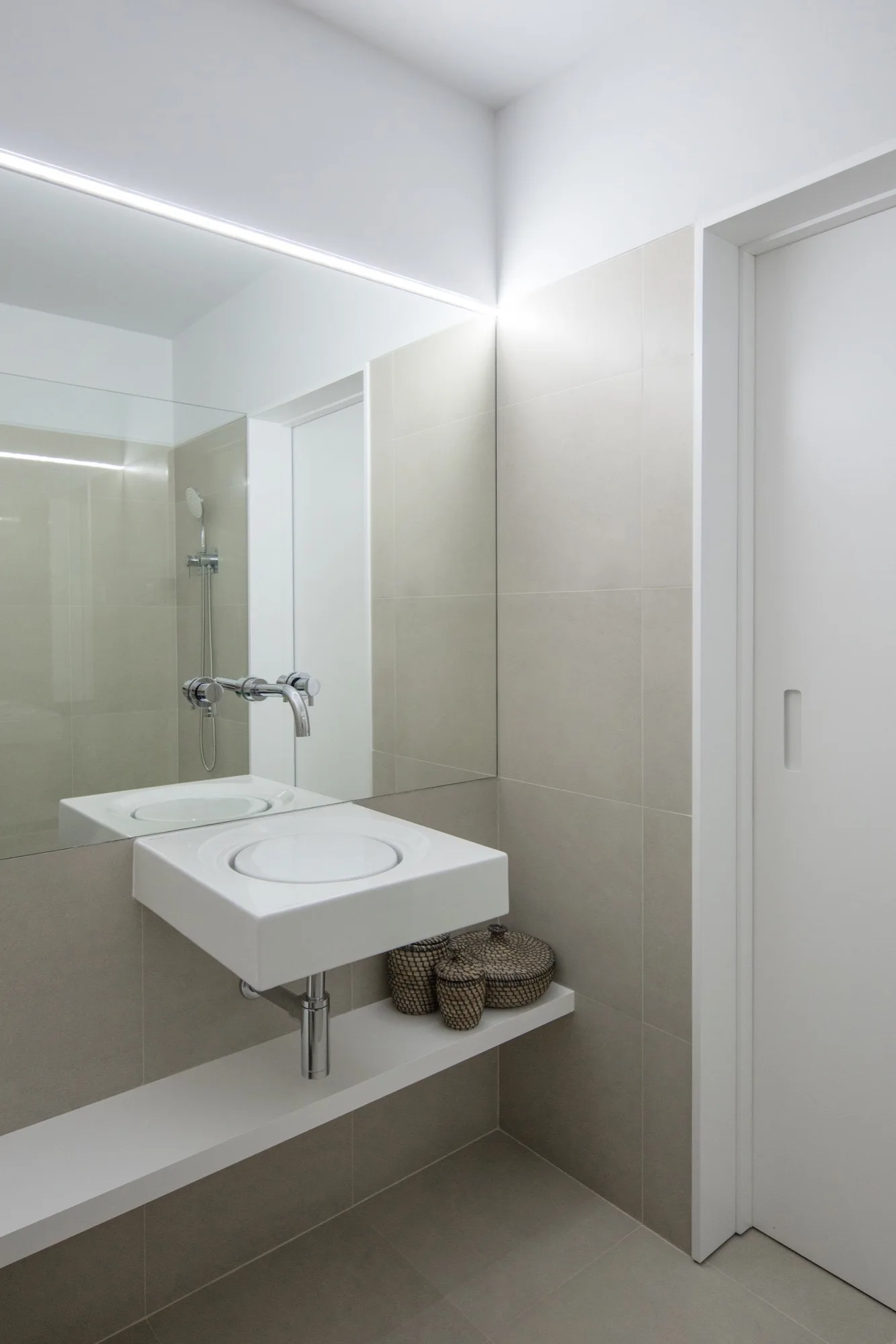 A minimalist bathroom features a white wall-mounted sink, a wall-mounted faucet set in a large mirror, and matte beige wall tiles.
