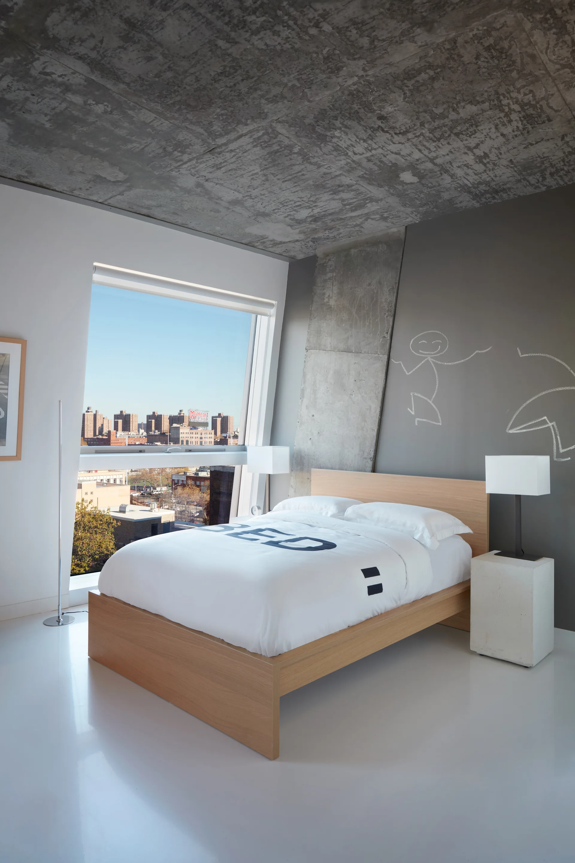 A modern bedroom features a light wood platform bed, an angled window overlooking a city, a glossy white floor, and an exposed concrete ceiling.