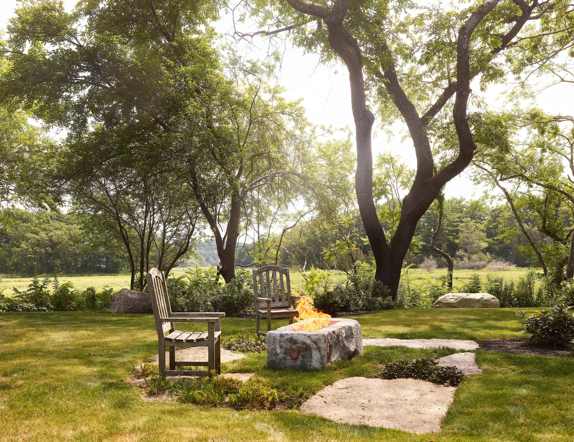 Two weathered wooden chairs stand beside a rustic stone fire pit with an active flame on a grassy lawn with stone pavers.