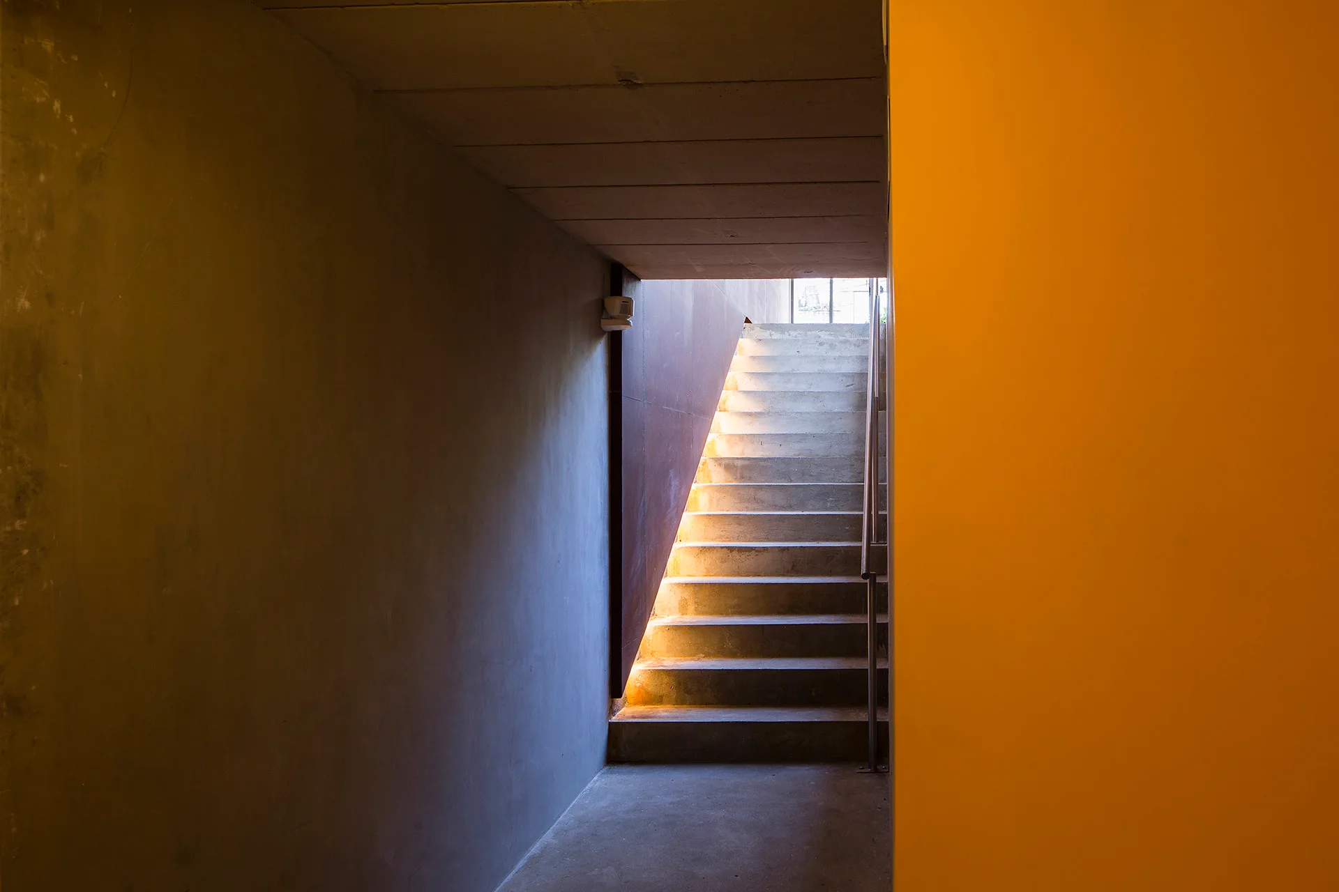 A minimalist concrete staircase rises from a grey hallway next to a large orange wall and is lit by a warm side light strip.
