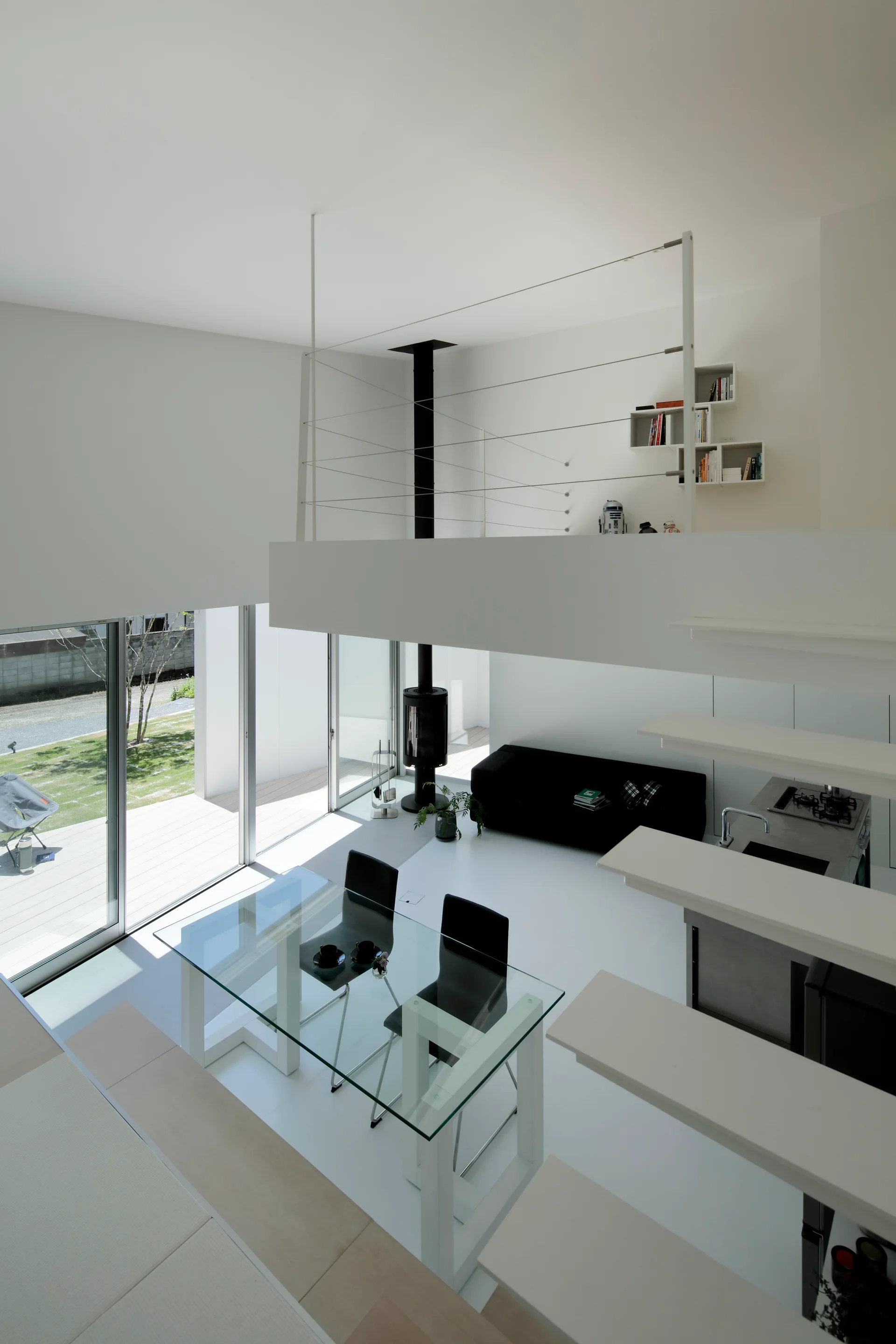A minimalist double-height white living room with a glass dining table, floating stairs, a black sofa, and a black wood-burning stove.