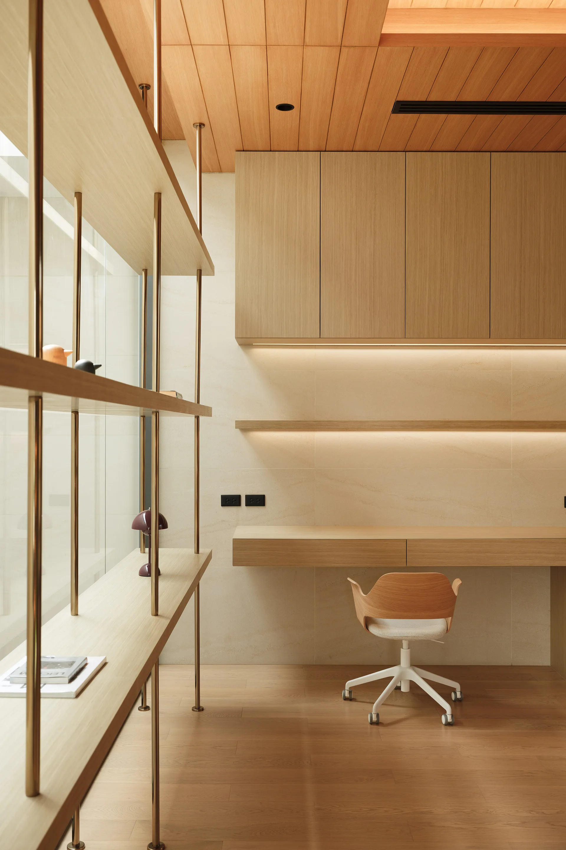 A minimalist home office features a floating wooden desk, a matching modern chair, upper wood cabinets, and a tall shelf with brass supports.