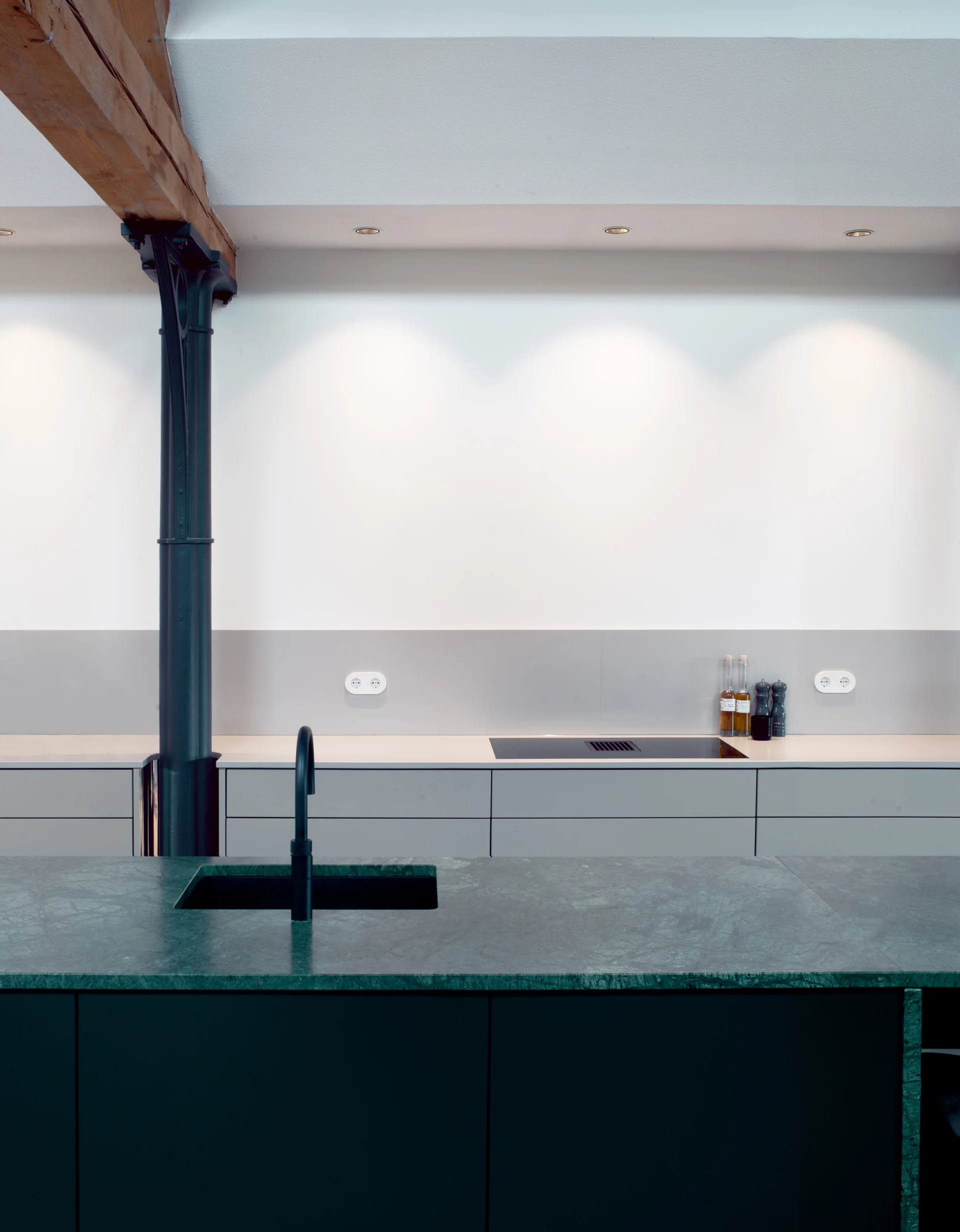 A minimalist kitchen featuring a dark green marble island with a black faucet, gray cabinetry, and an industrial support column.