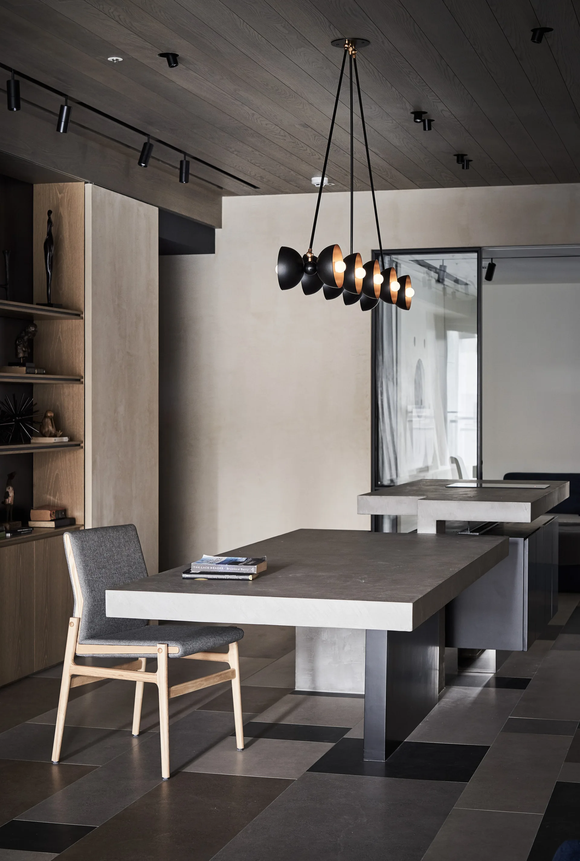 A minimalist kitchen features a split-level concrete island, a grey dining chair, and a black multi-arm pendant light below a dark wood ceiling.