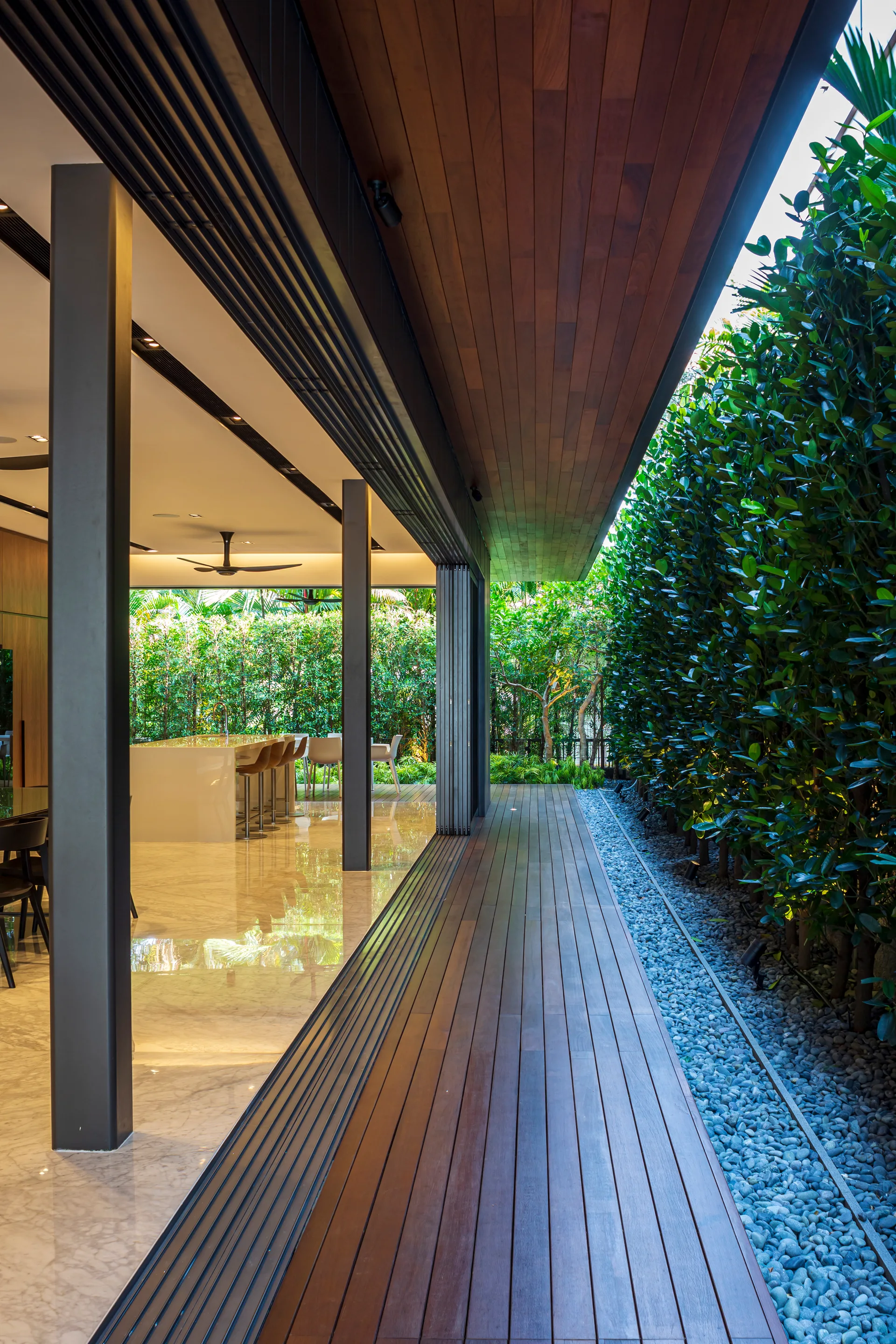A modern wooden terrace deck features a matching wood ceiling, a pebble border with a tall green hedge, and open sliding doors showing a kitchen interior.