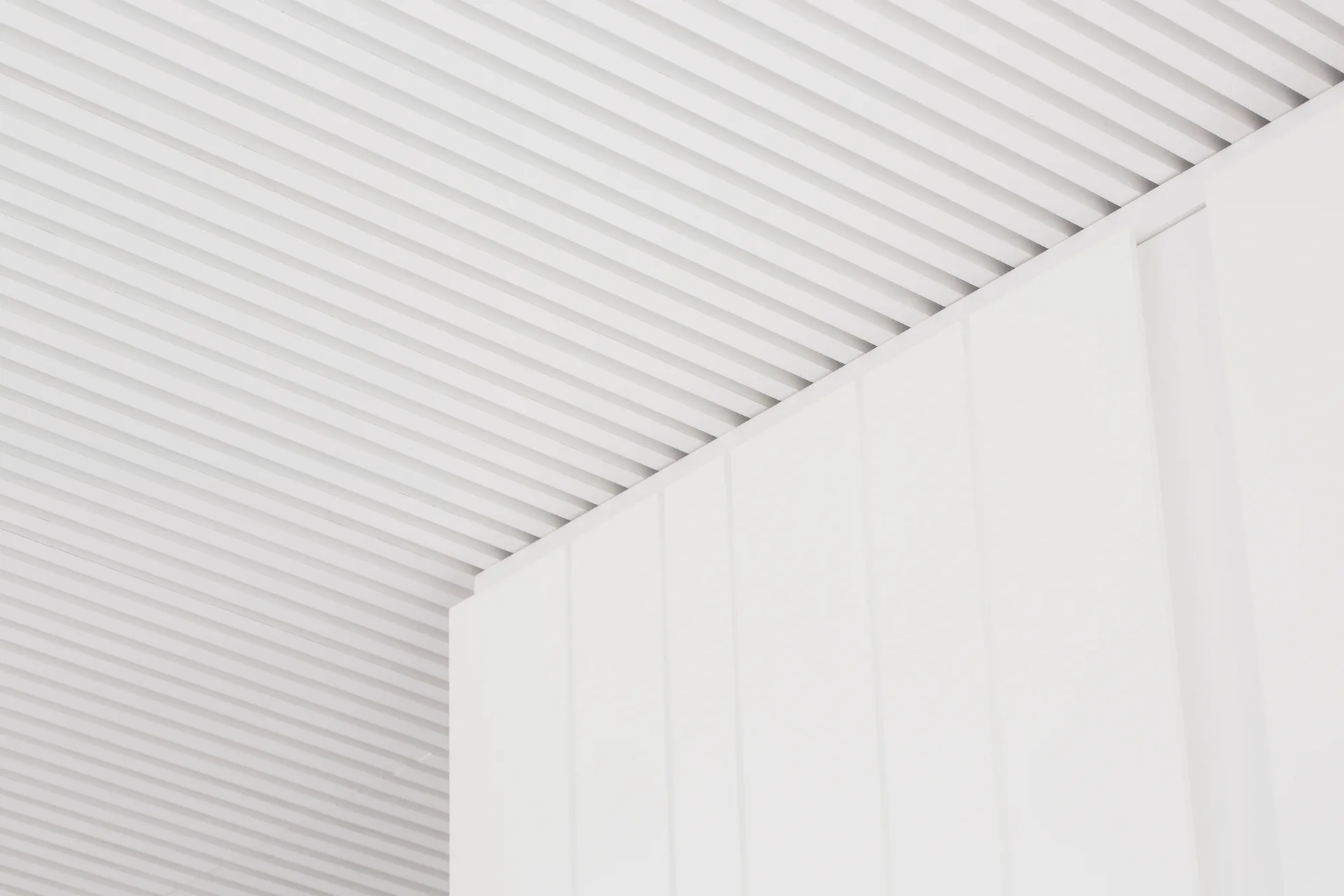 A close-up shot of a minimalist white slatted ceiling meeting a smooth, vertically paneled white wall.