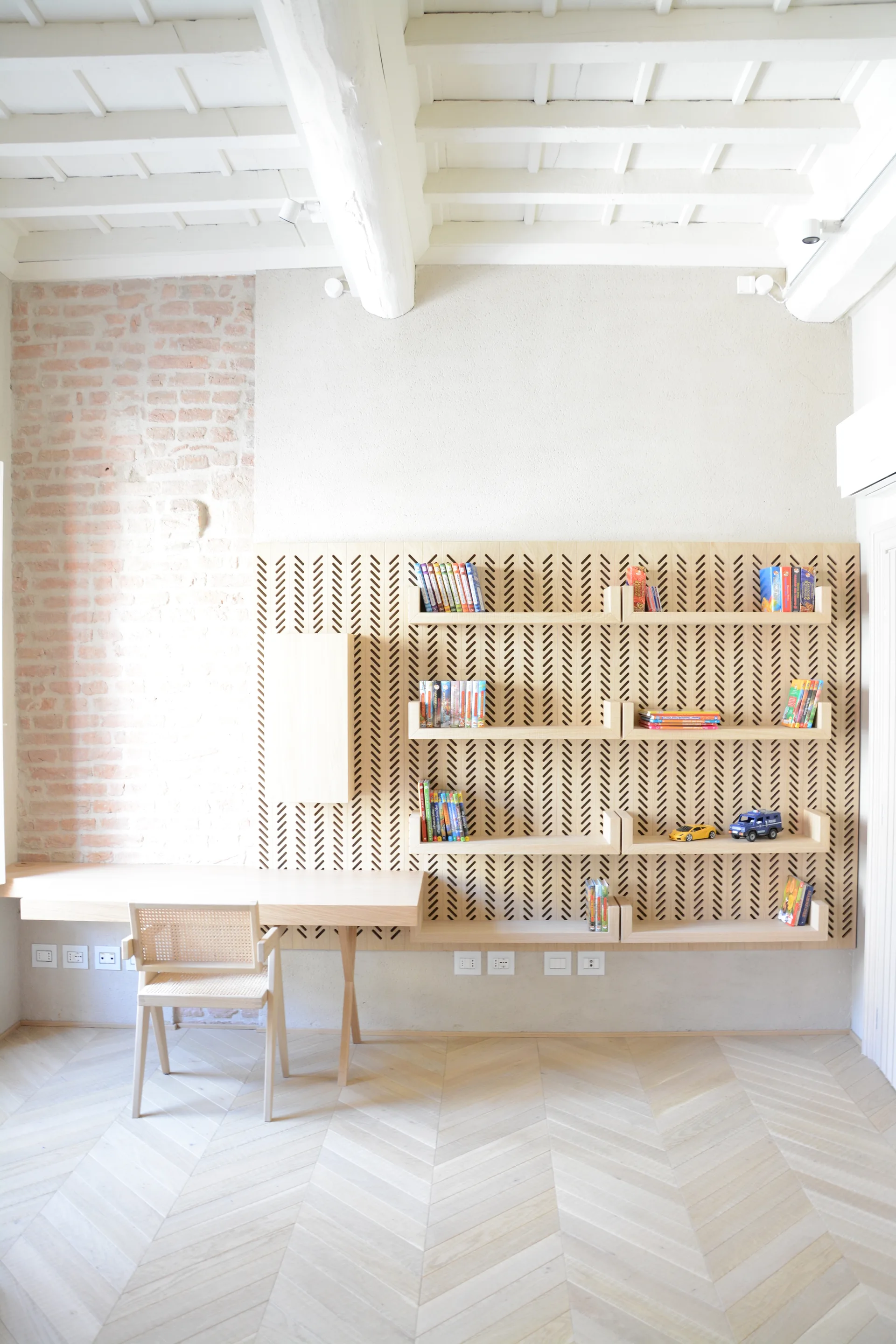 A bright home office features a light wood desk and chair, a slotted wooden wall panel with shelves, herringbone parquet flooring, and exposed brick.