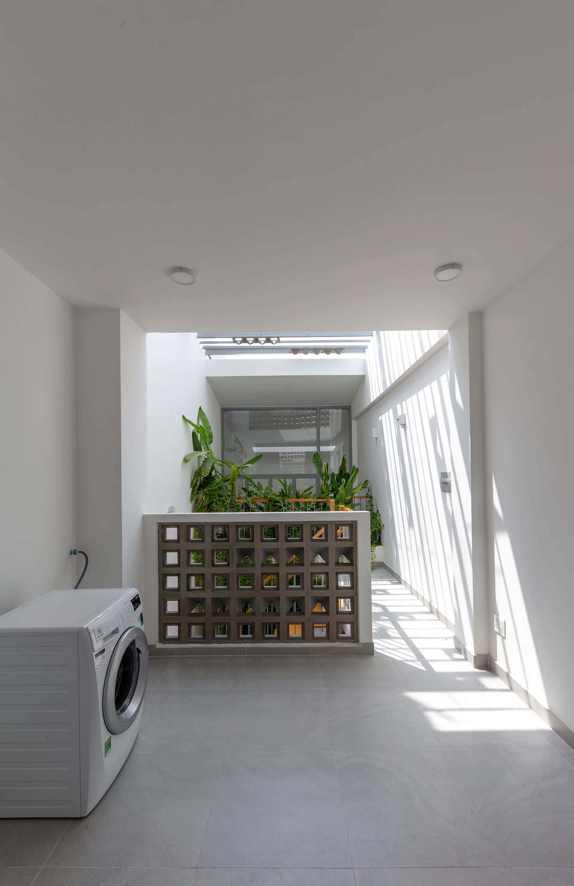 A minimalist utility space features a washing machine on grey tiled floors, a concrete breeze block partition, and plants under a skylight.
