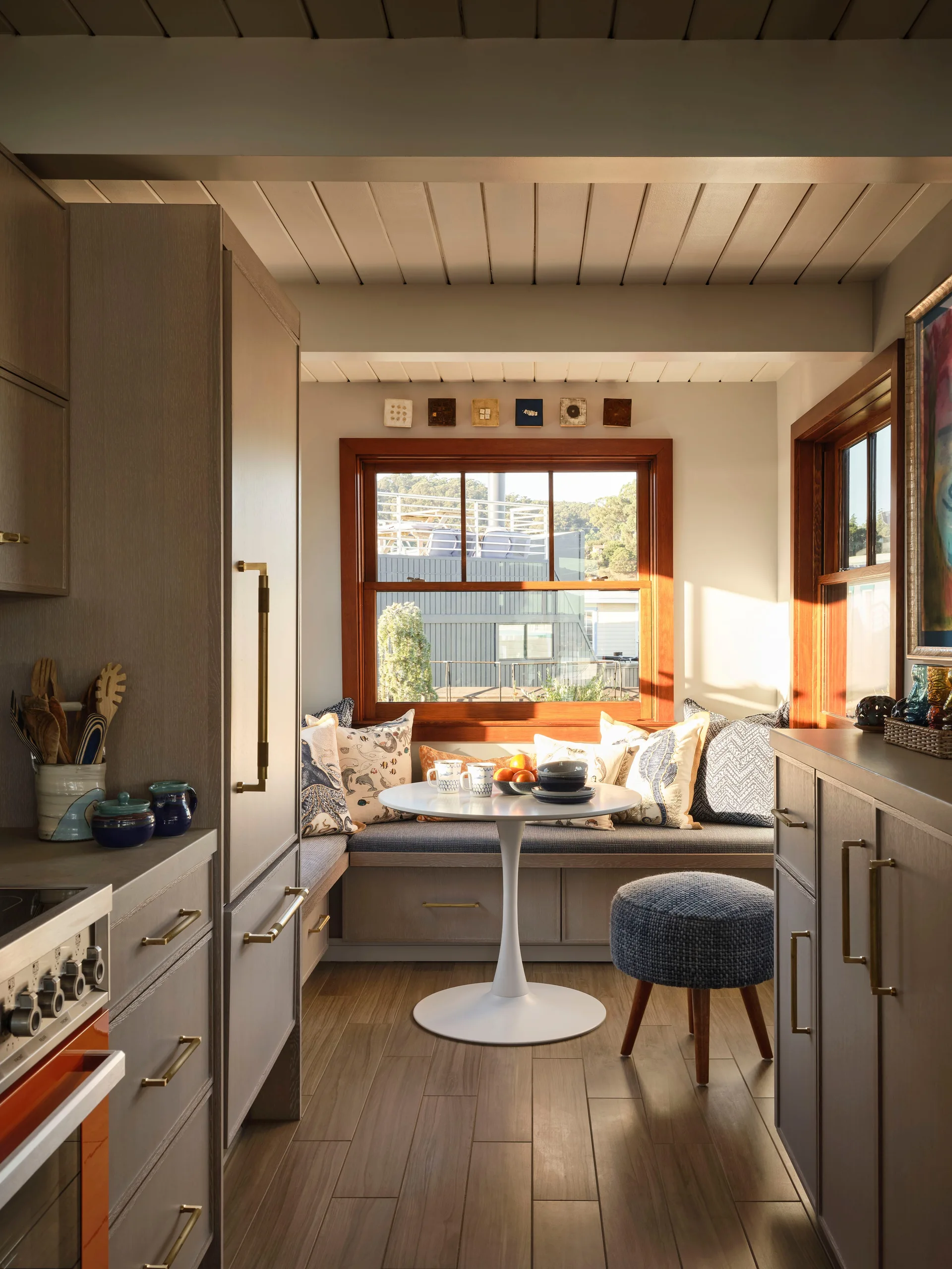 A sunlit kitchen breakfast nook features a built-in corner bench with patterned cushions, a white pedestal table, and grey cabinetry.