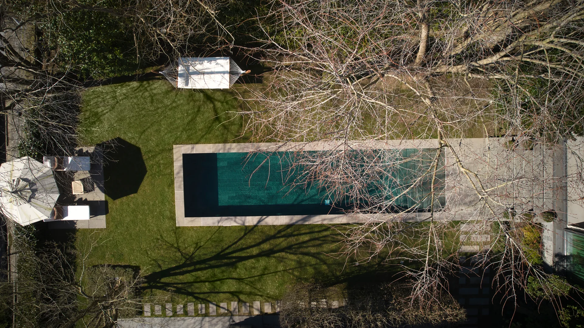 An aerial view of a rectangular swimming pool surrounded by a green lawn, stone stepping stones, lounge chairs, and bare trees.