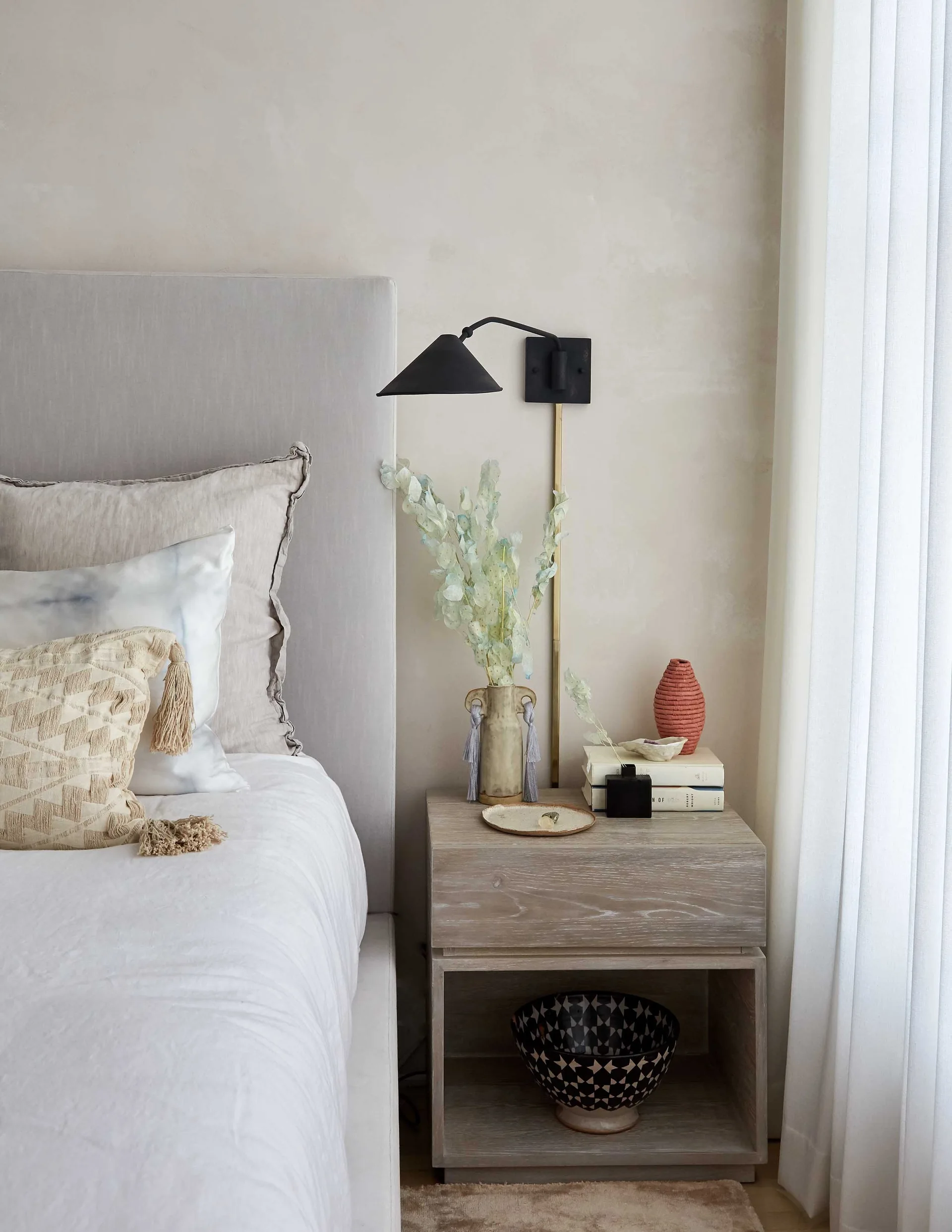 A light wood nightstand sits beside an upholstered bed, featuring a black metal wall sconce, a vase with leaves, and a textured tassel pillow.